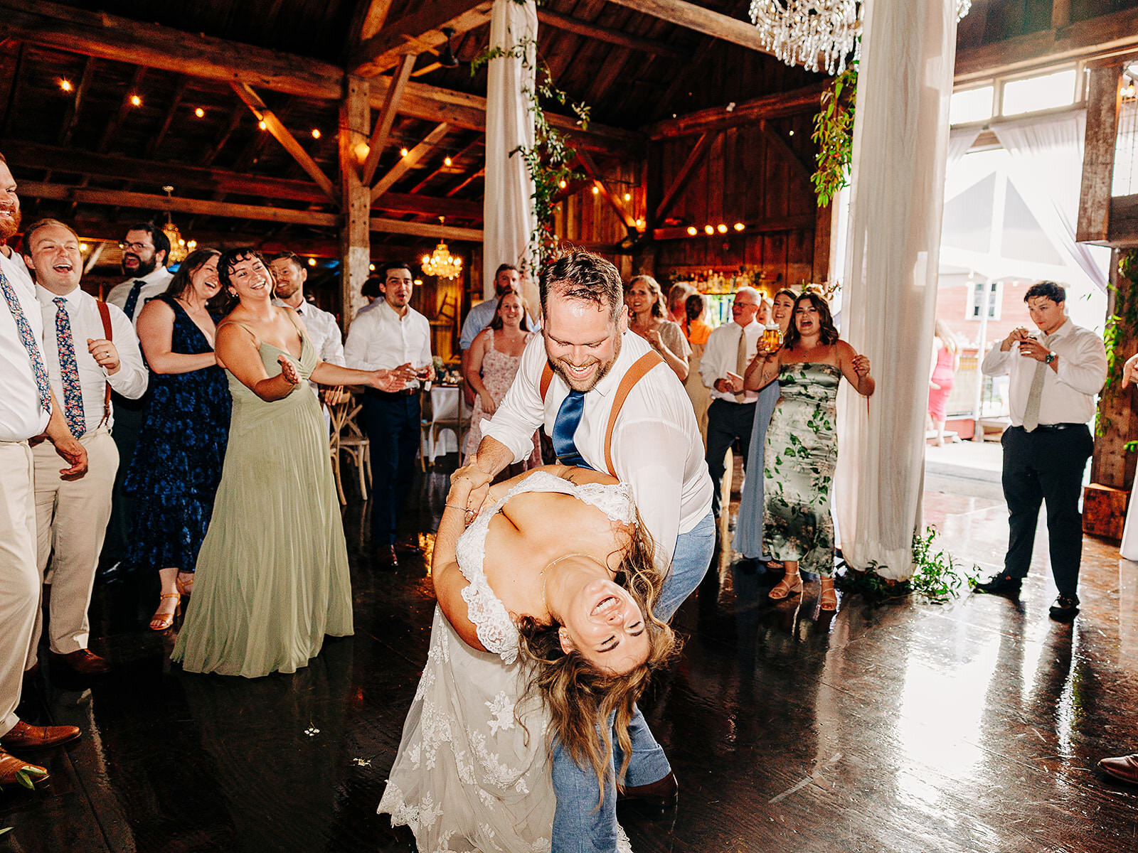 A groom joyfully dips his laughing bride on the dance floor as friends and family cheer around them in a rustic, warmly lit barn during a wedding celebration in Eau Claire WI. 