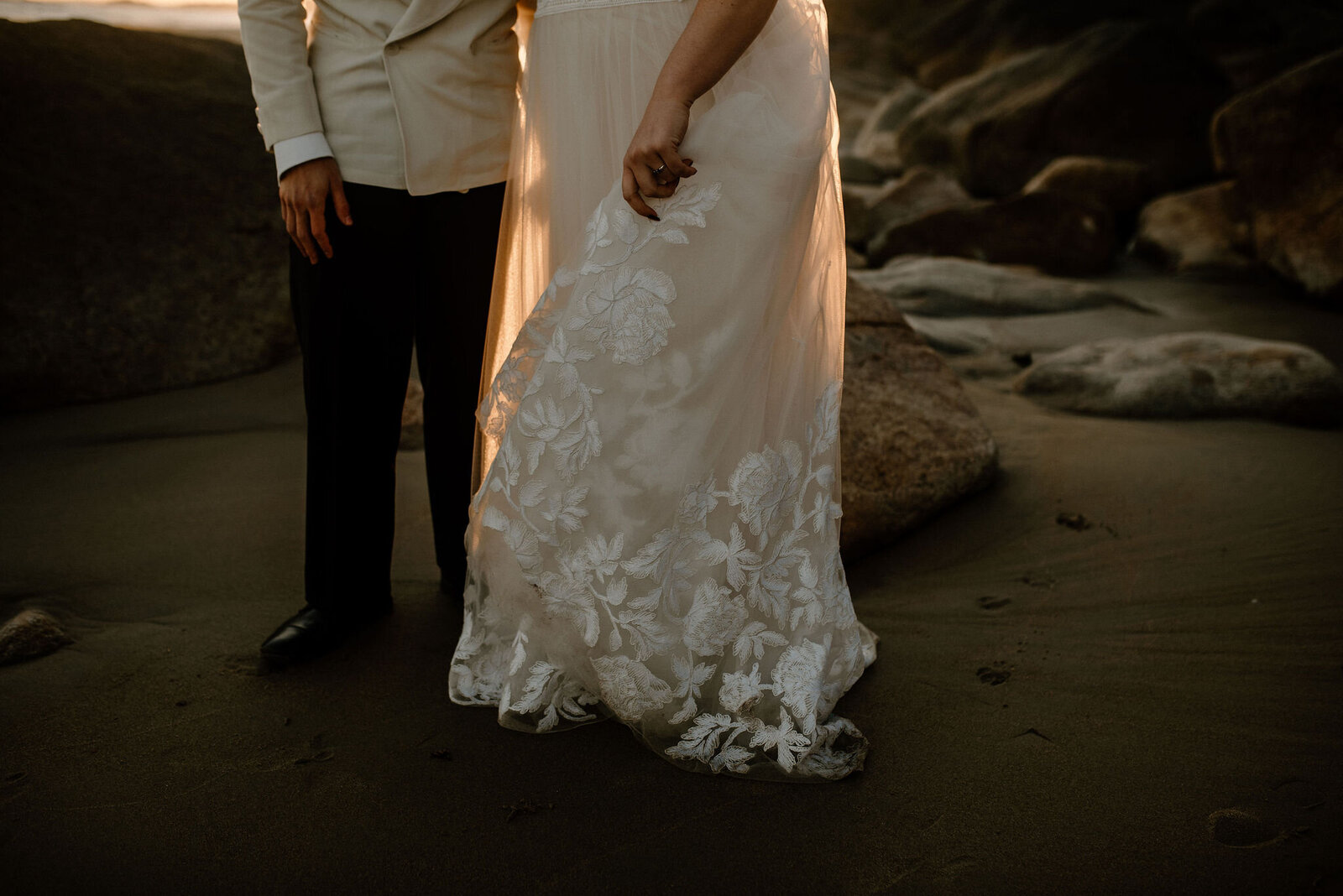 couple standing on rocky beach at sunset featuring lace details on bottom of wedding dress