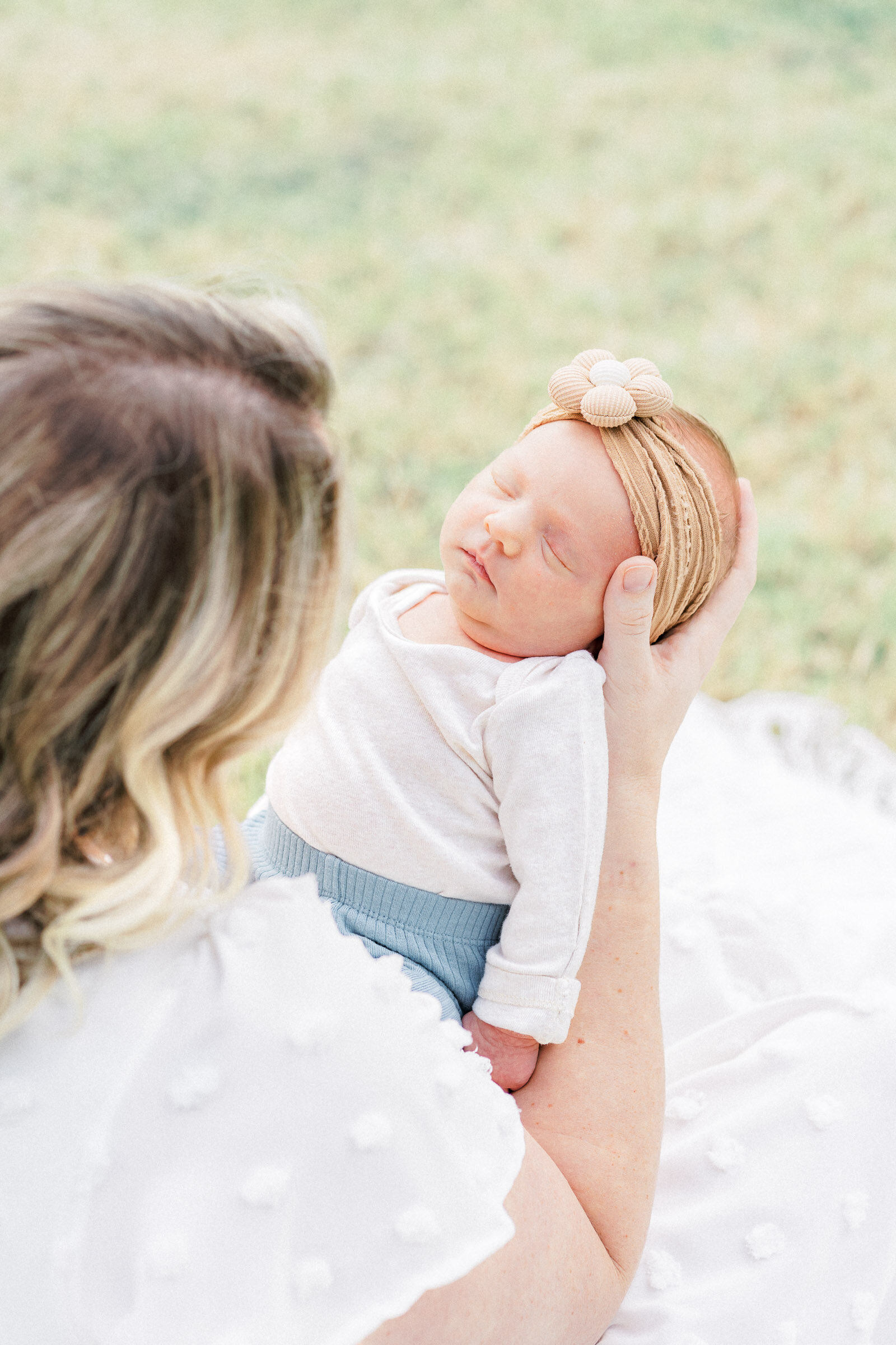 a woman dressed in a long white dress sits on on the grass and holds her baby on her lap facing her. The baby is sleeping.