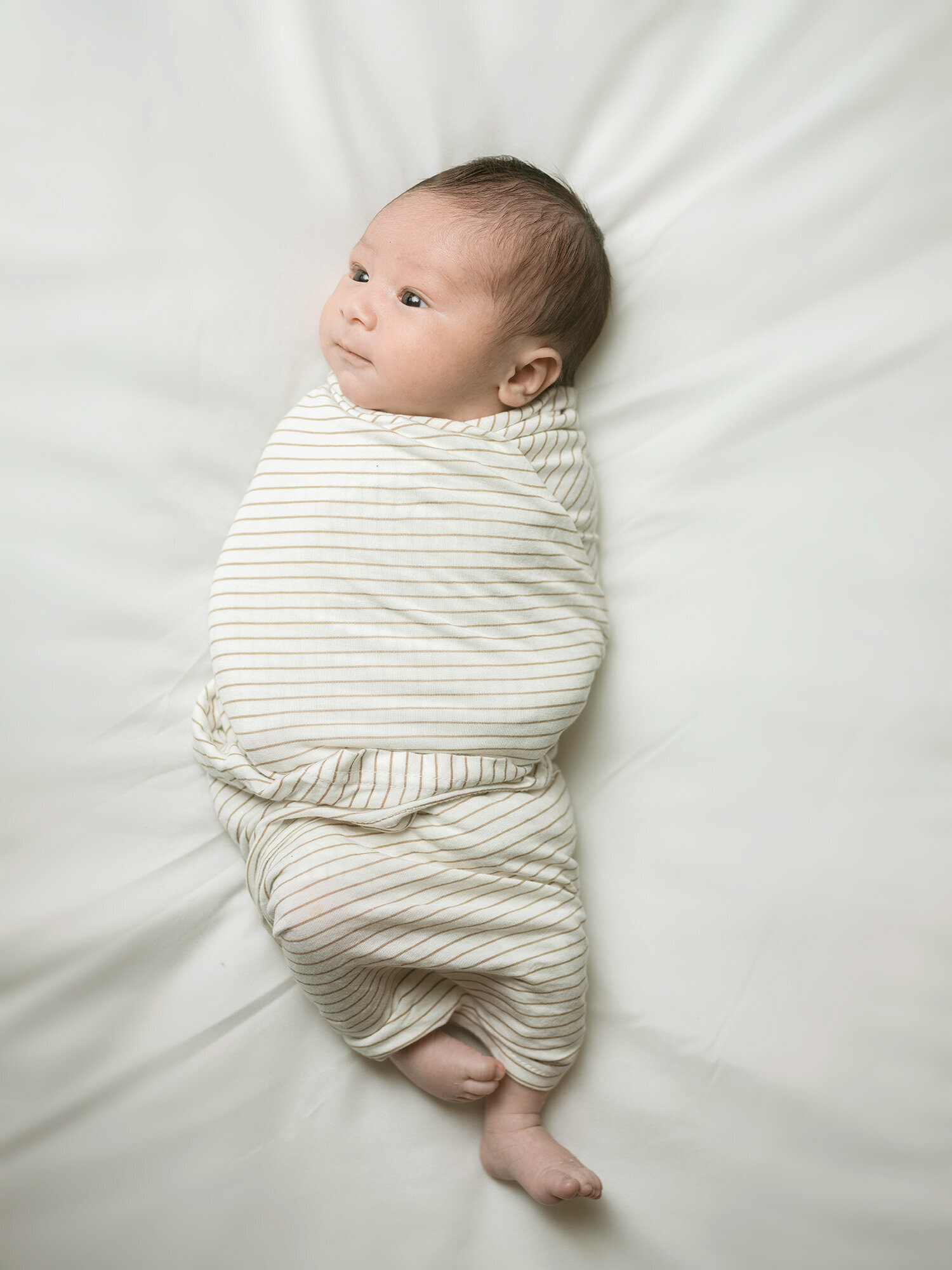 Newborn baby wrapped in a striped swaddle on white bedding, photographed during an in-home newborn session by a Los Altos newborn photographer