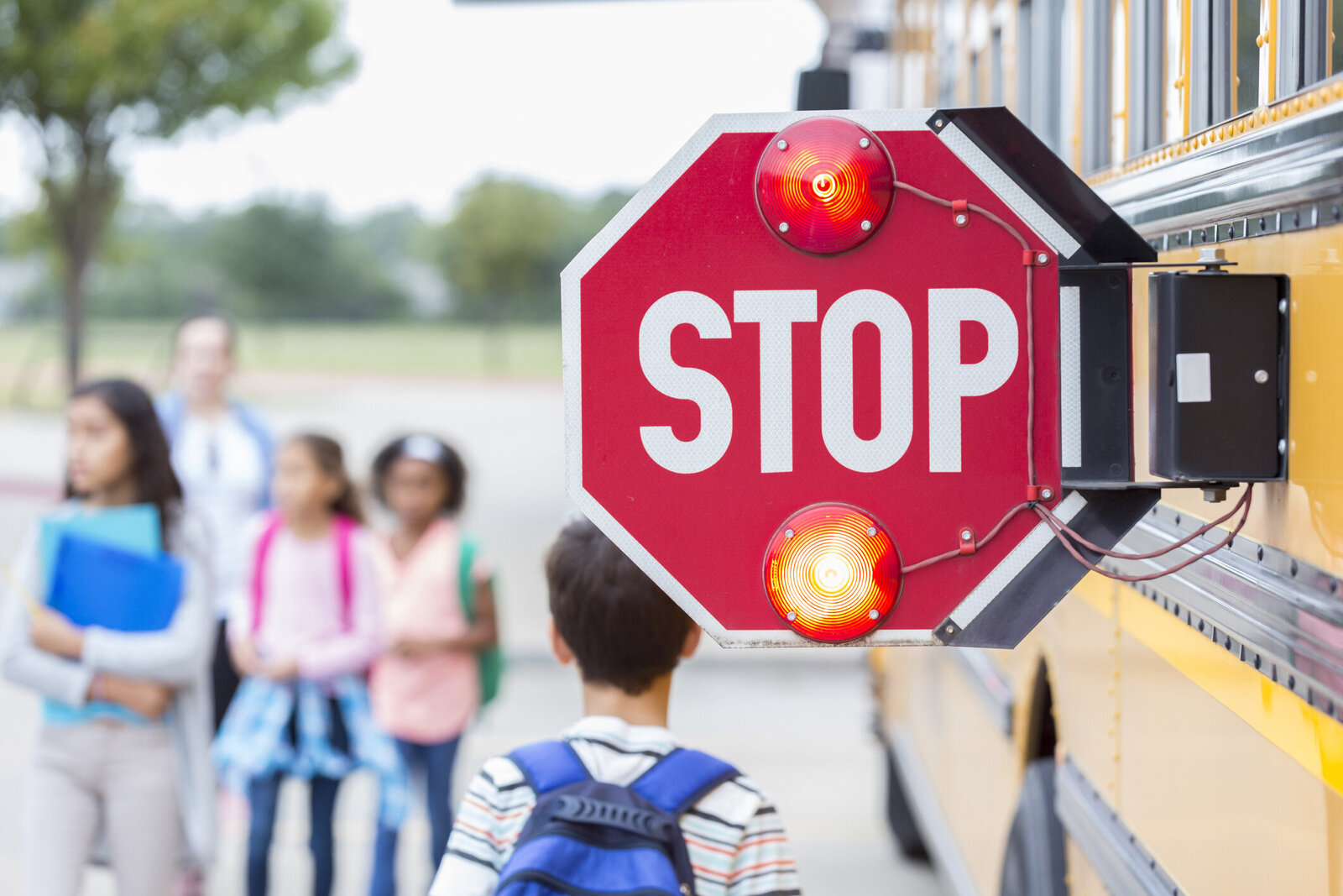 A close up view of a school bus stop sign  with a blurred view of children waiting  to get on in the background