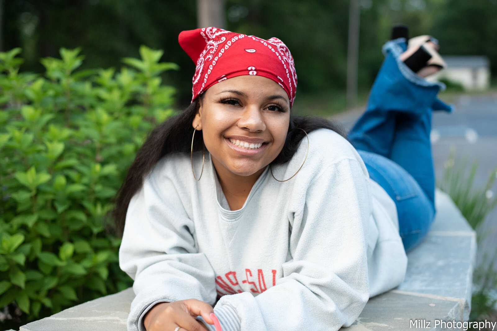 a senior wearing a white sweater with a red bandana on her head laying with her feet crossed behind her smiling at the camera as she poses for her graduation photos photographed by Millz Photography in Greenville, SC