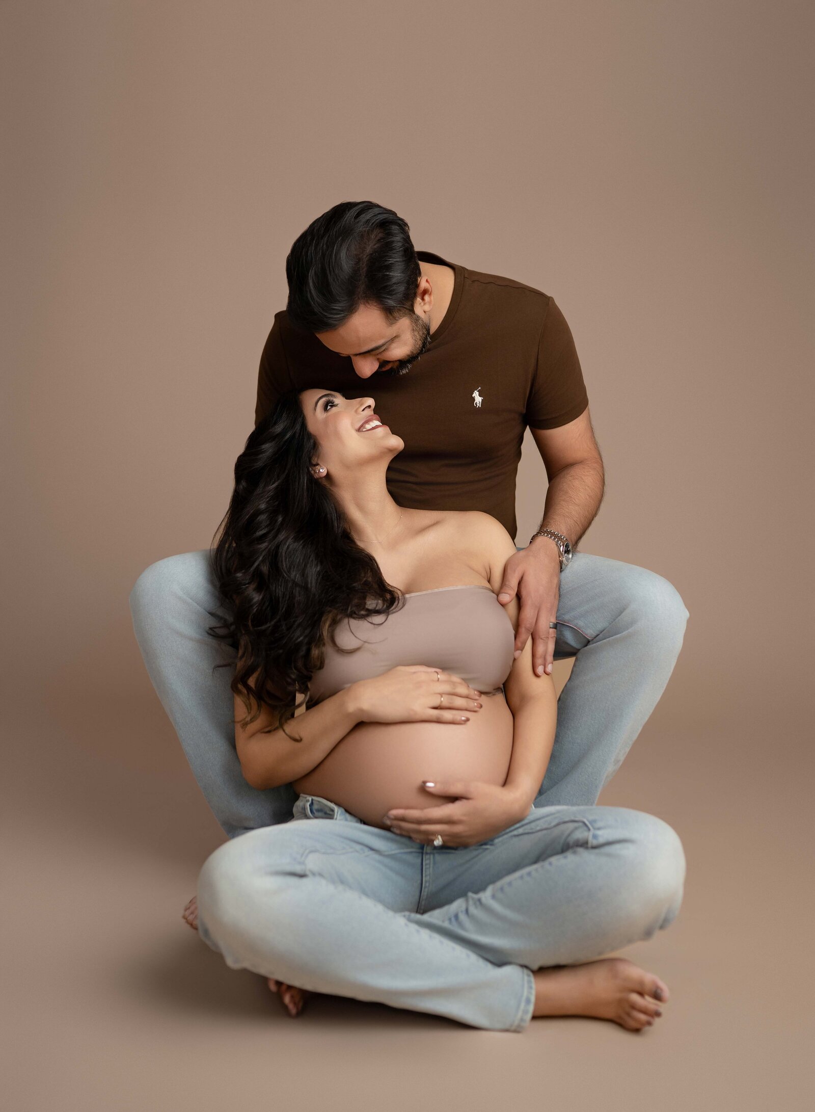 Mom sitting between Dad's legs on tan backdrop with baby bump out and smiling at each other. Photo taken at Atlanta Maternity photo studio in Woodstock, GA.