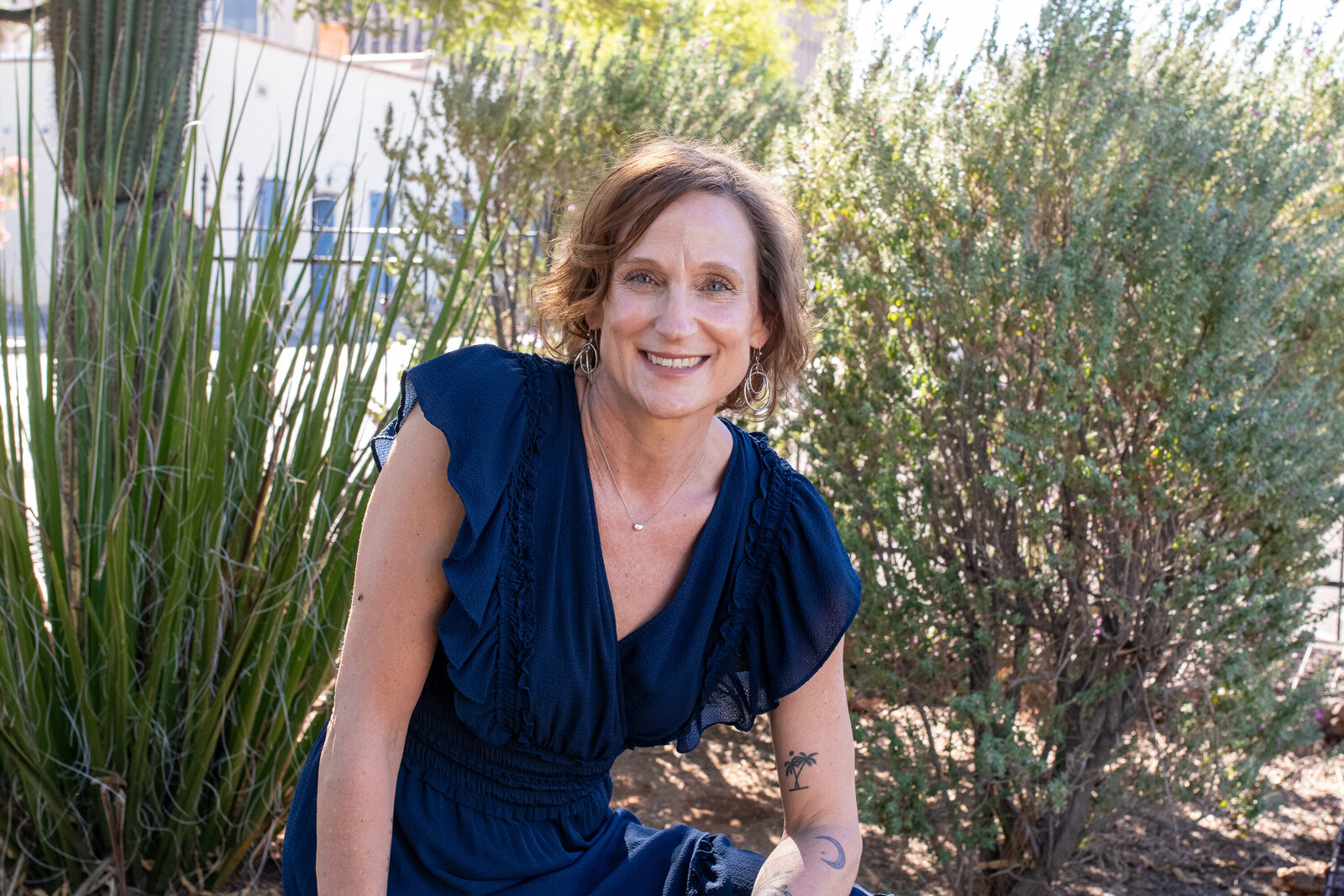 Smiling woman in a blue dress seated among tall desert plants outdoors, photographed by Vyrl Photo, highlighting Tucson brand photography.