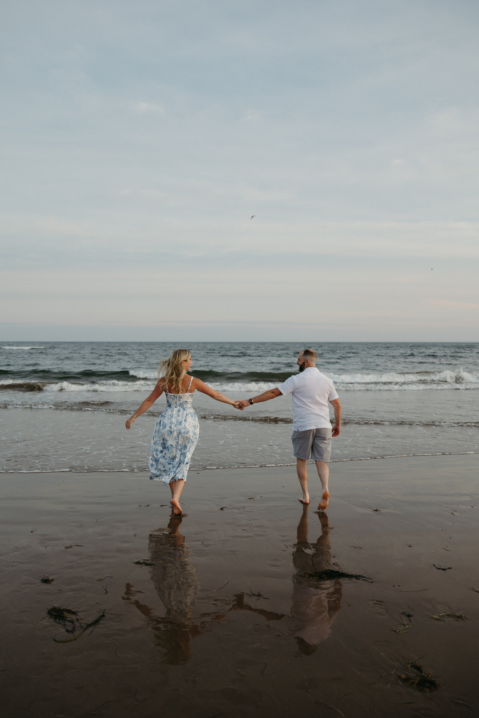A couple holds hands and runs towards the ocean after getting engaged at Conrad's beach in Halifax.