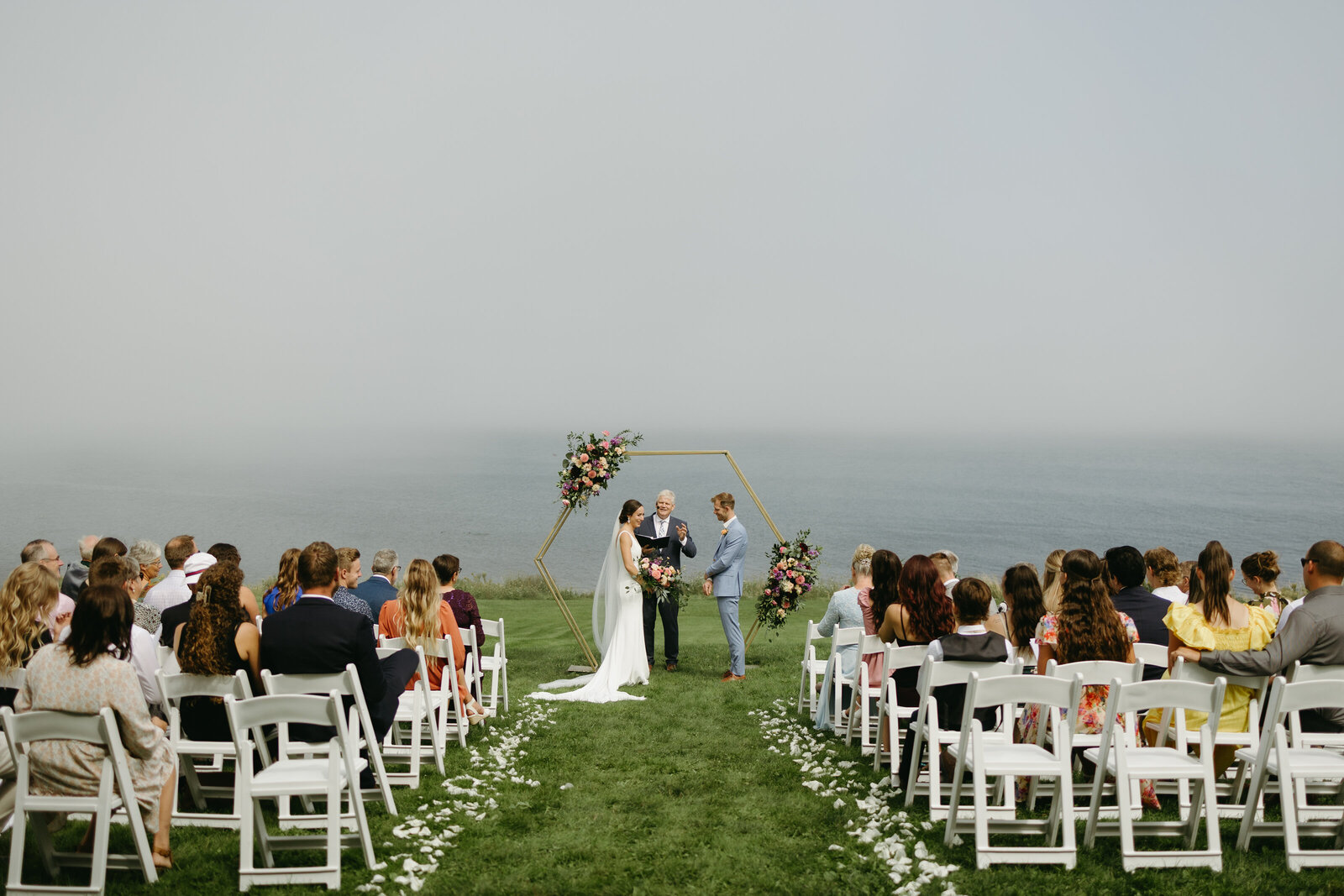 A small intimate coastal ceremony where a bride and groom are getting married and are being watched by their closest family and friends. 