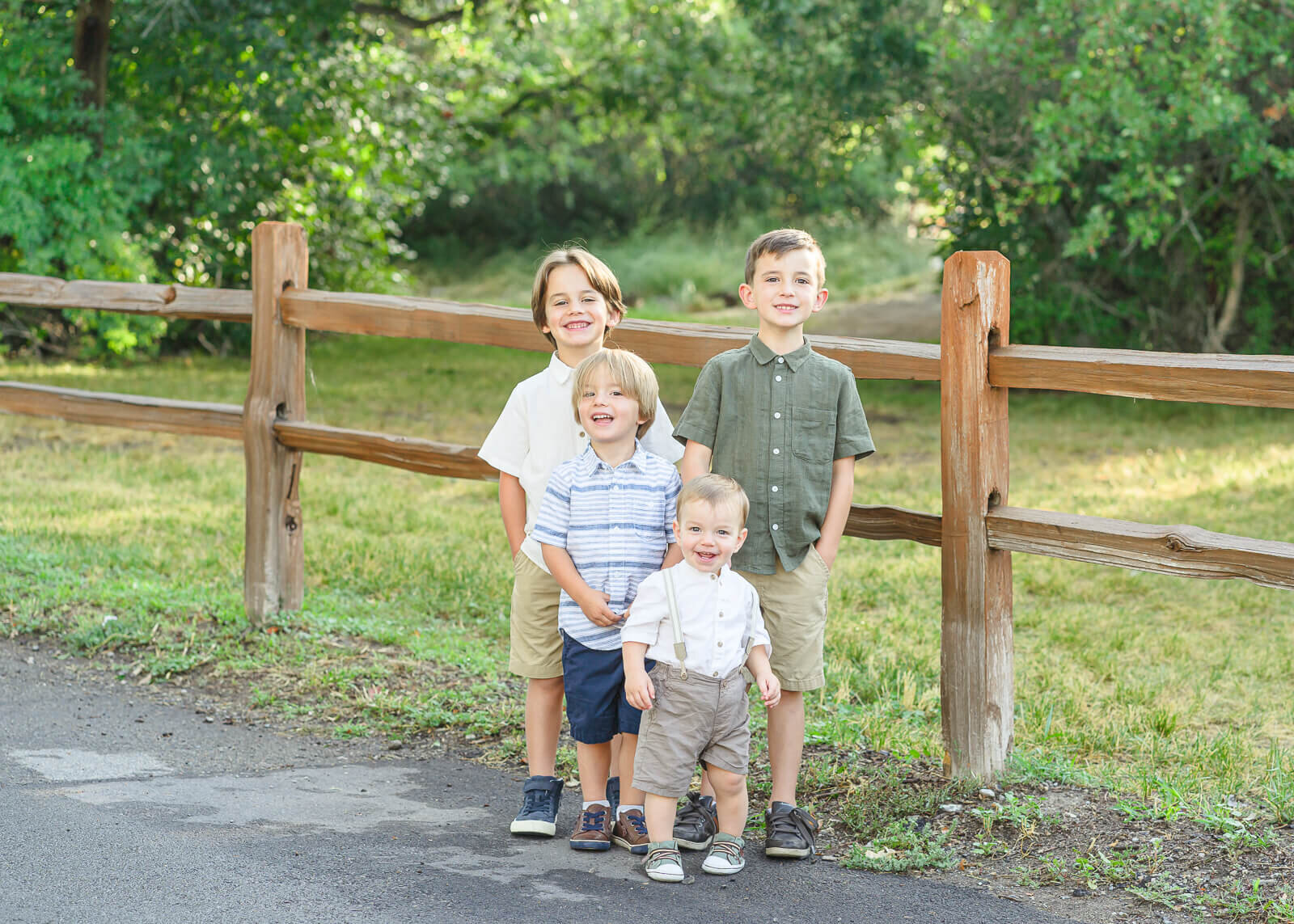 Four young boys standing in front of a wooden fence with sunlight filtering through the green oak trees in the background. Captured at Highland Glen Park.