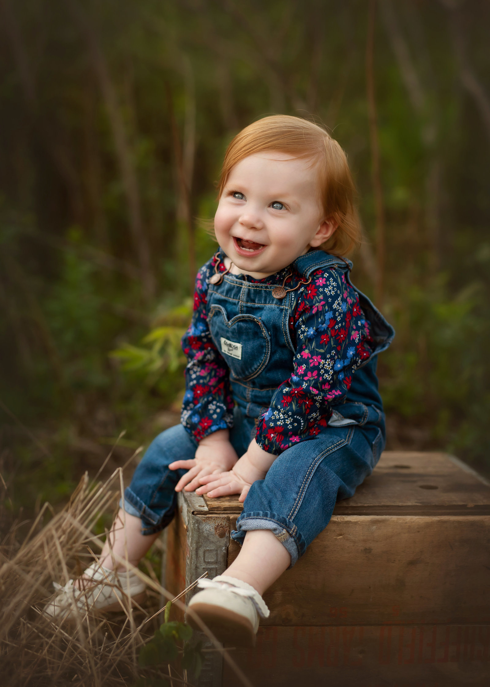 red headed baby in overalls smiling while sitting on a wooden box outside