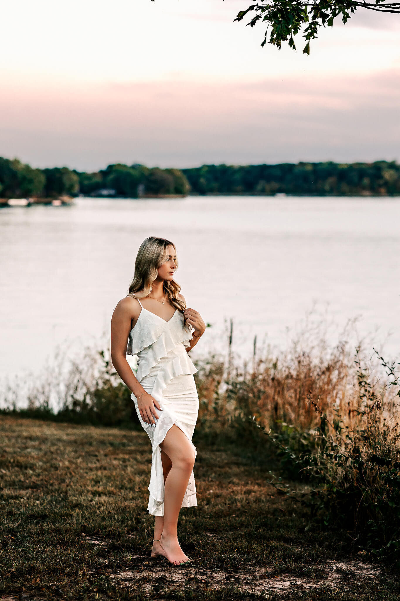 A woman in a white dress stands on grass near a lake during sunset. She gazes to the side, with trees and a serene water view in the background. The sky is softly colored with pink and orange hues.