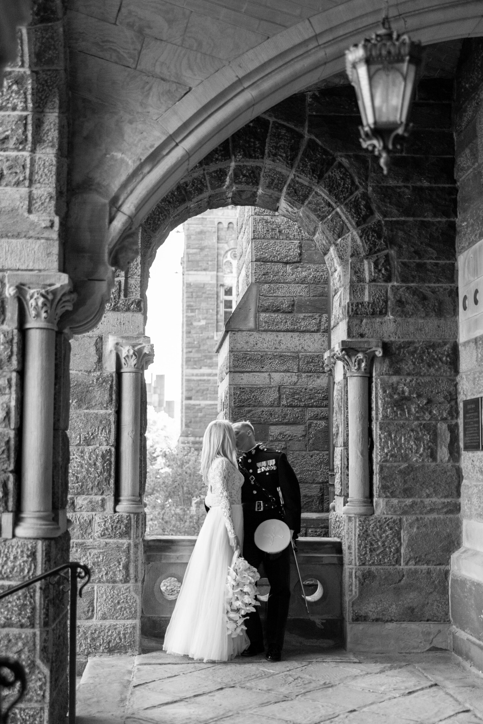 A bride and groom share a kiss under a stone archway. The bride holds a bouquet and wears a long gown, while the groom is in a formal military uniform. The setting is Gothic with detailed stone architecture.
