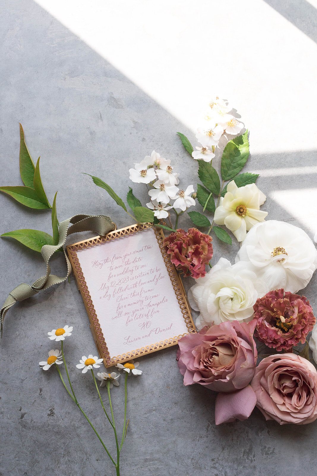 Flat lay photograph of wedding details including floral accents such as white ranunculus, daisies, lisianthus, and mauve garden rose.