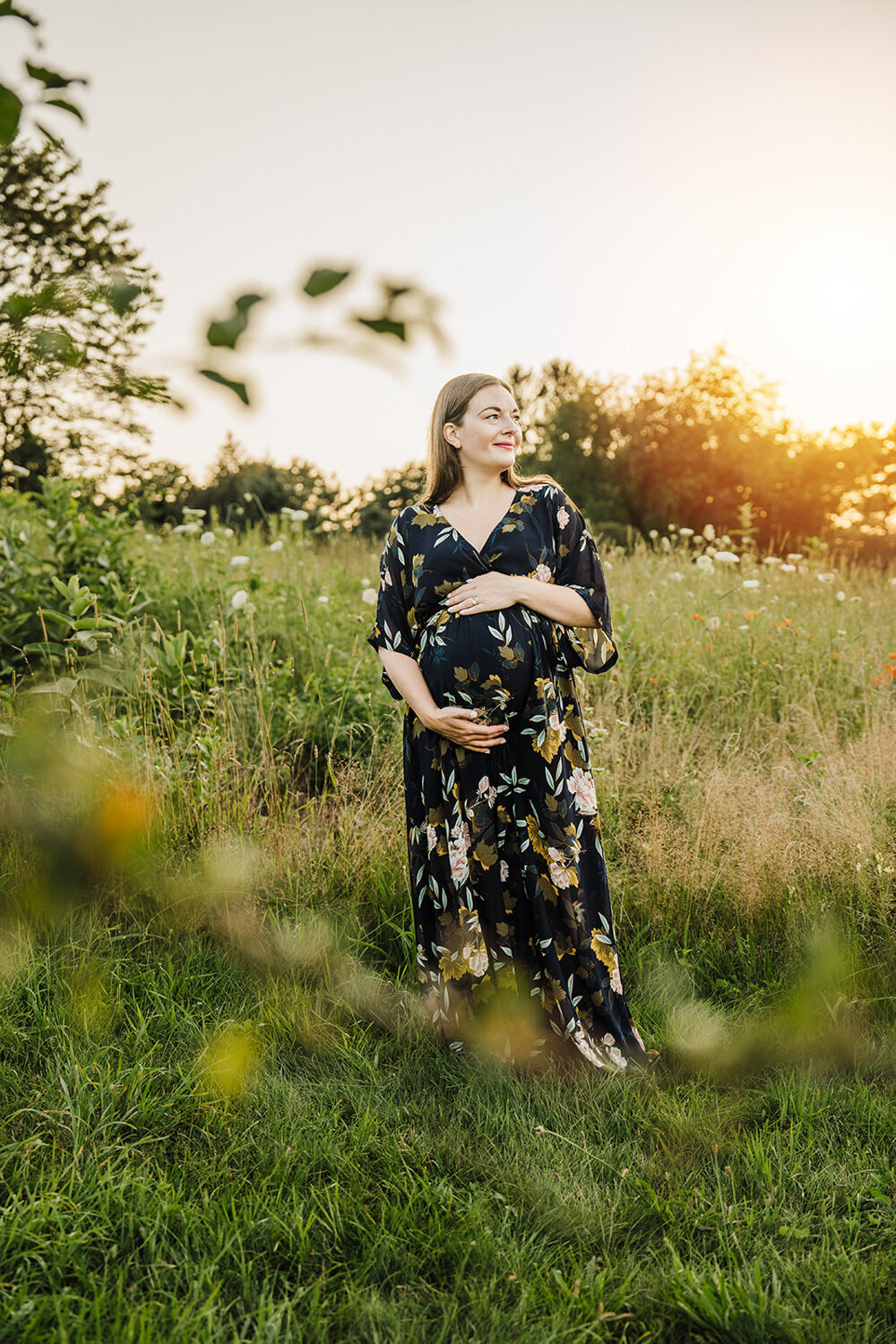 pregnant mom in floral dress stands in a field at sunset