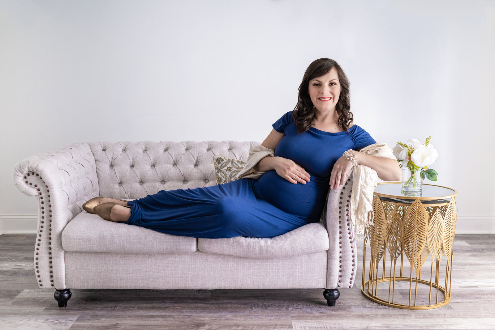 A pregnant woman lays across the sofa and poses for her maternity portraits in the studio