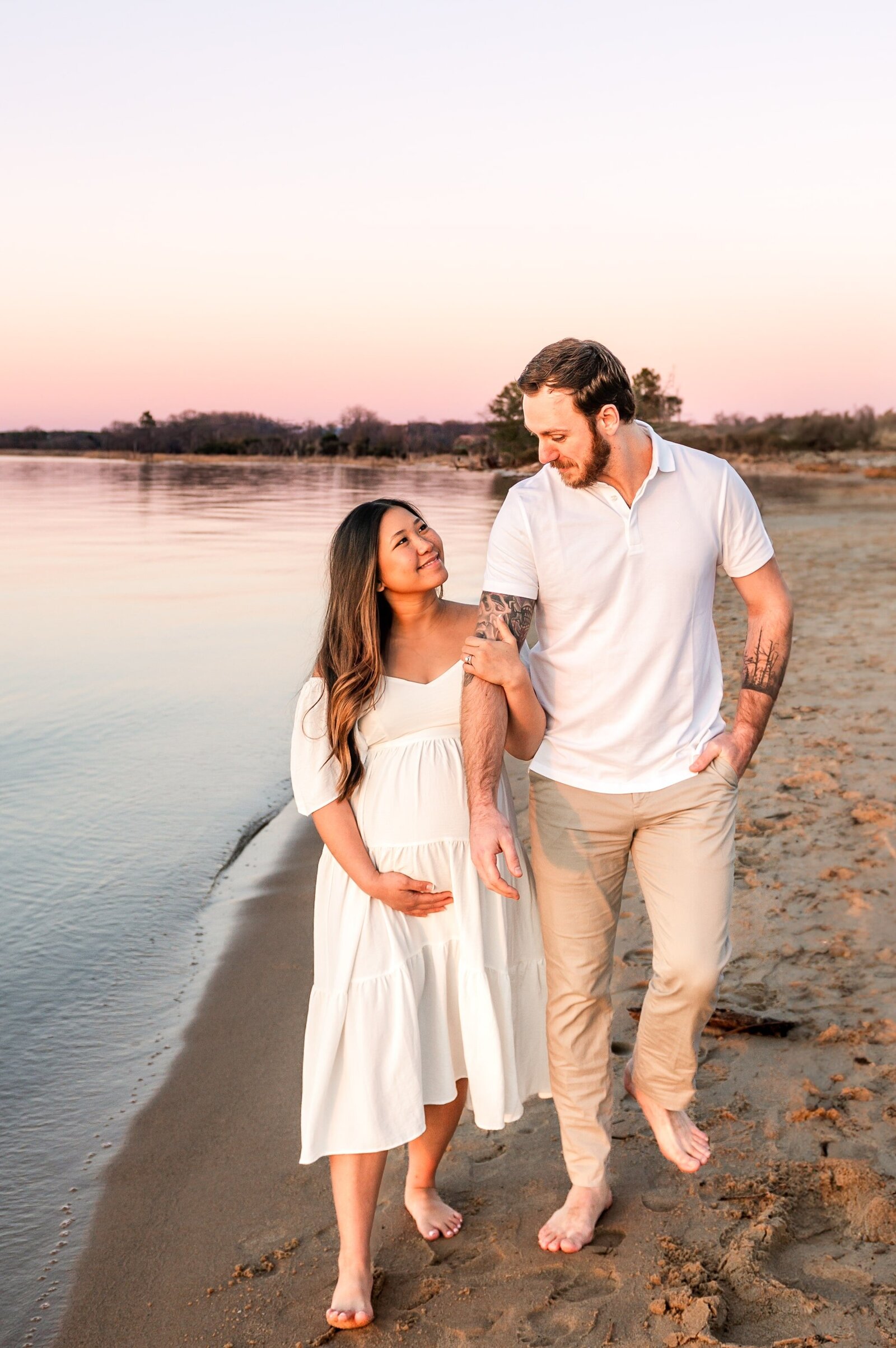 pregnant woman and husband walk along Annapolis beach during sunset