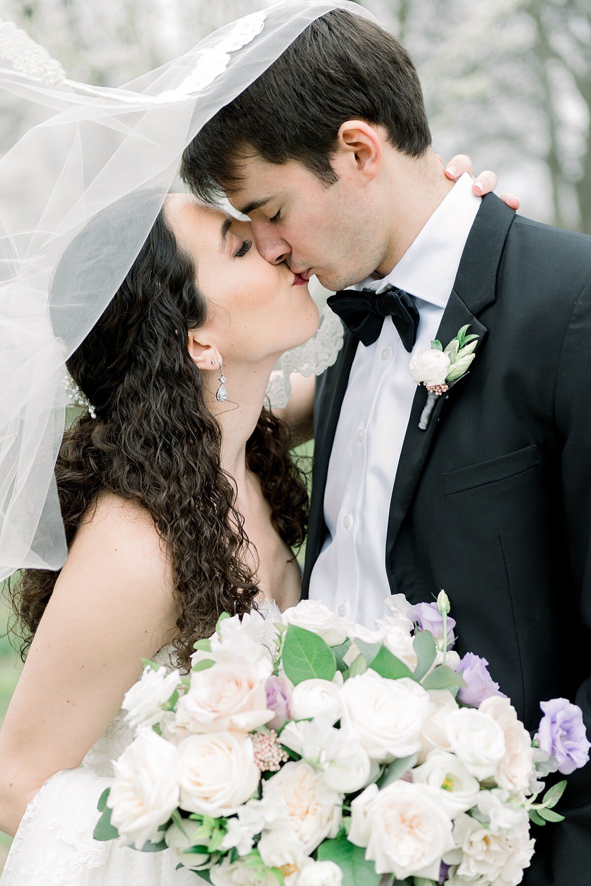 Bride and groom kissing underneath the bride's veil while bride holds large bouquet of white and purple flowers