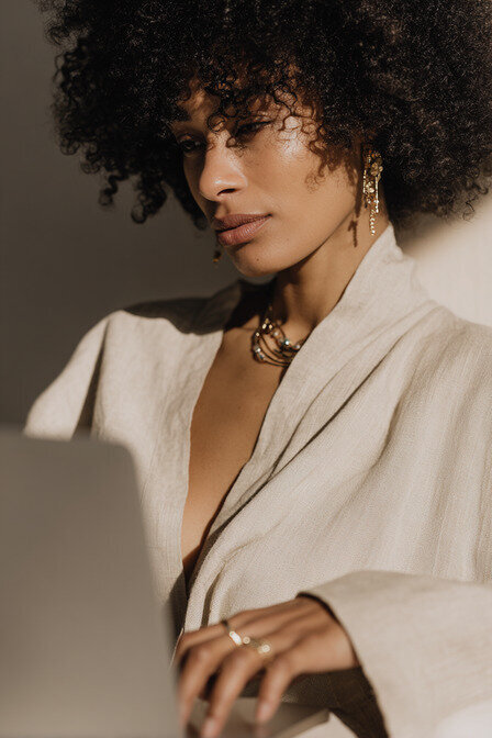 Editorial portrait of a designer working on a laptop in warm sunlight, wearing a neutral linen robe, representing the intentional and elegant aesthetic of Kathalyst Design Studio.