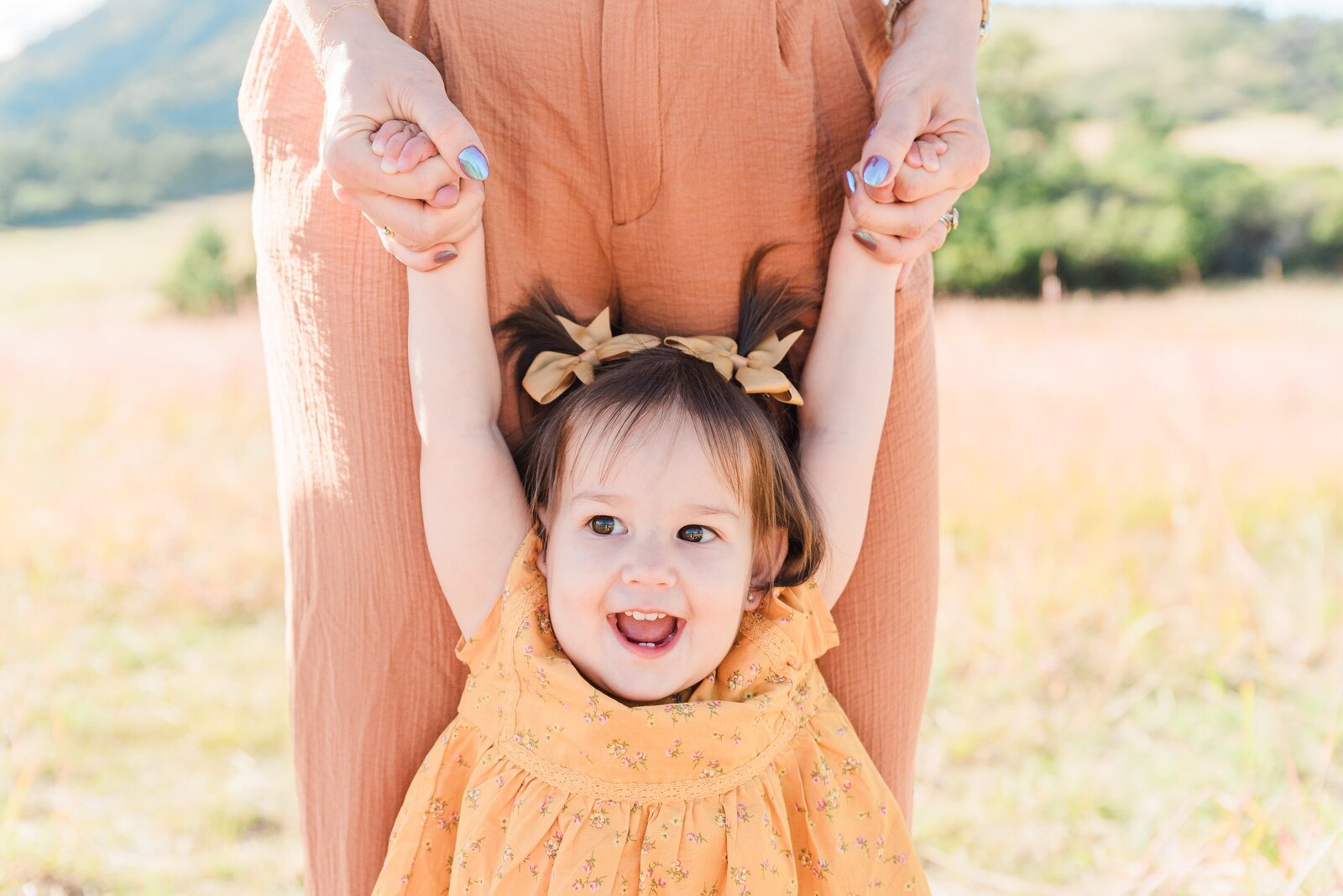 A woman in an orange jumpsuit holds a toddler in a yellow outfit in front of her legs for a close up shot of the toddler's face with her arms above her head captured by Denver family photographer