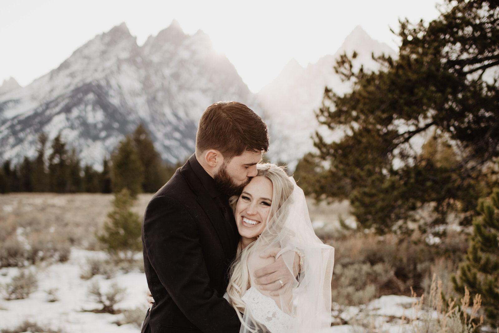winter wedding in the Grand Tetons captured by photographers Jackson Hole with groom hugging his bride and kissing her on the forehead as she beams with a smile and the sunsets over the mountains in the distance