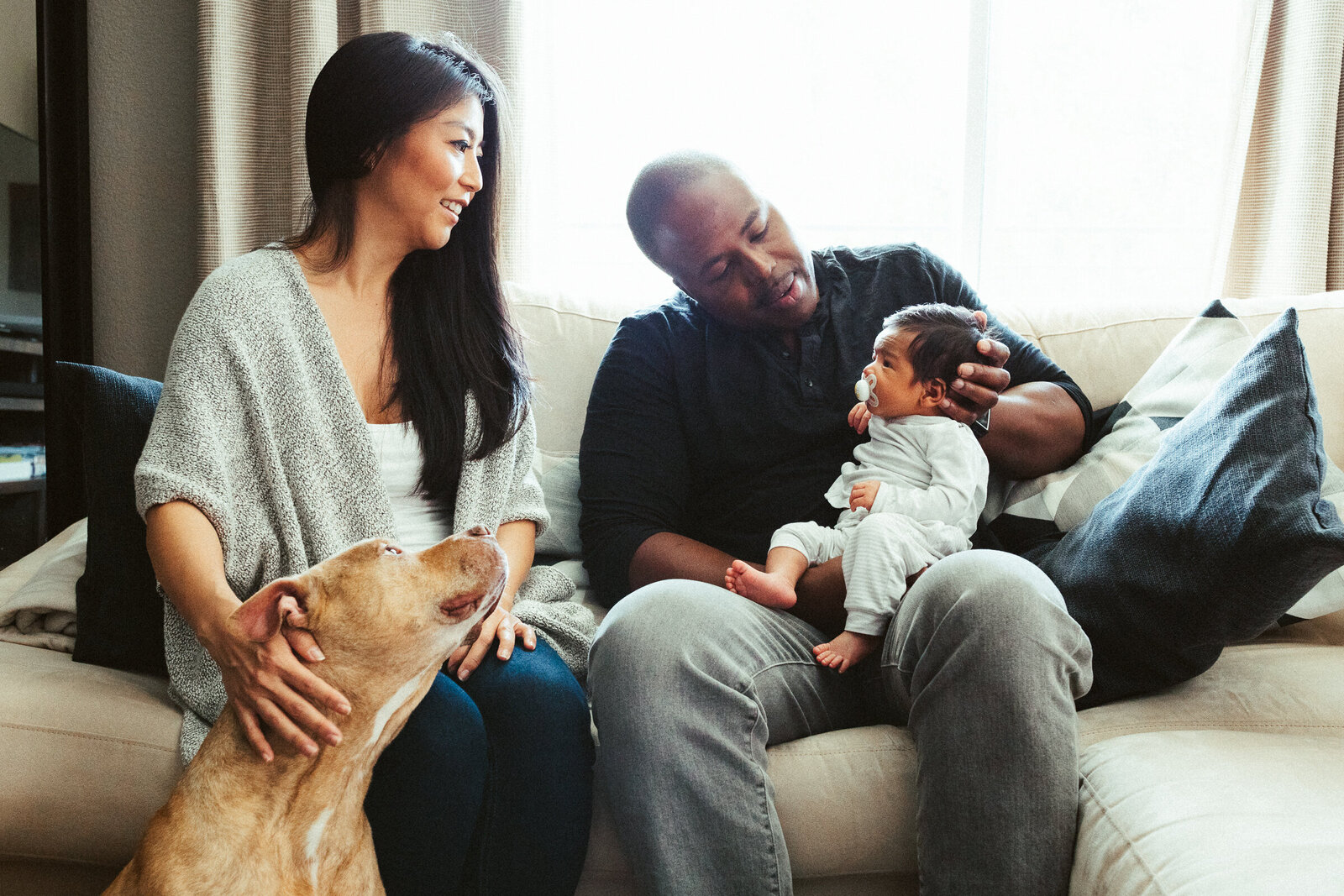 Asian mom and Black dad sitting in their living room holding their newborn baby boy while their dog looks on