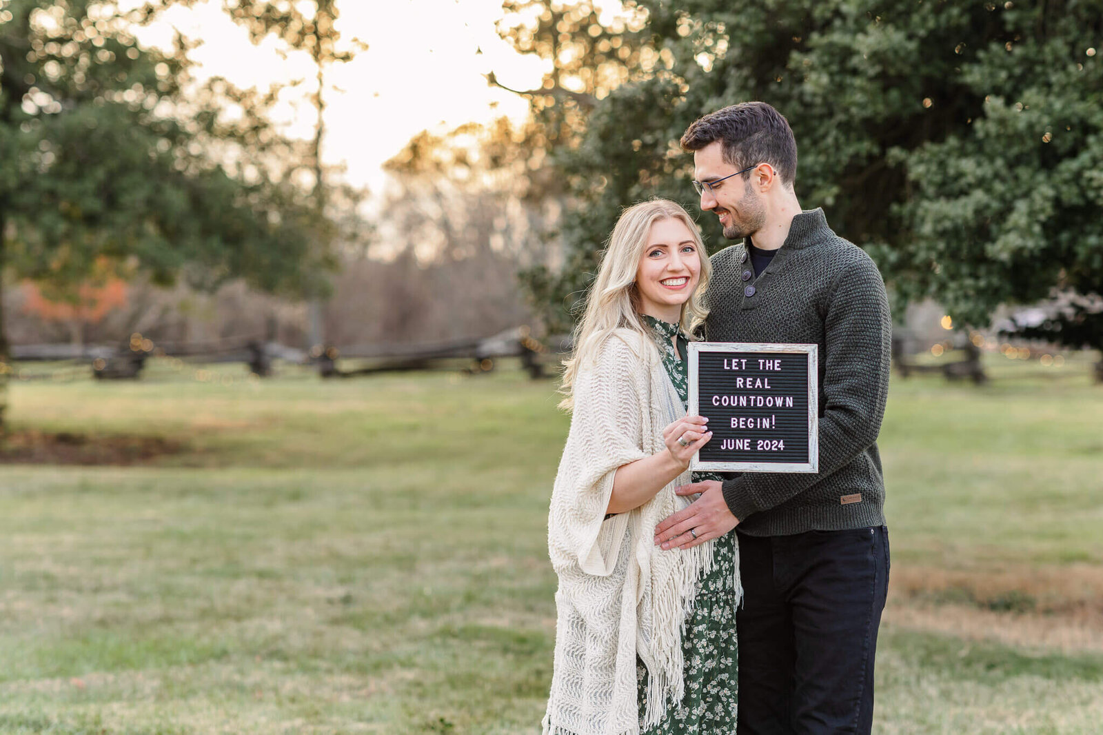 A mother and father to be snuggled up at a park at sunset holding a sign announcing their pregnancy.