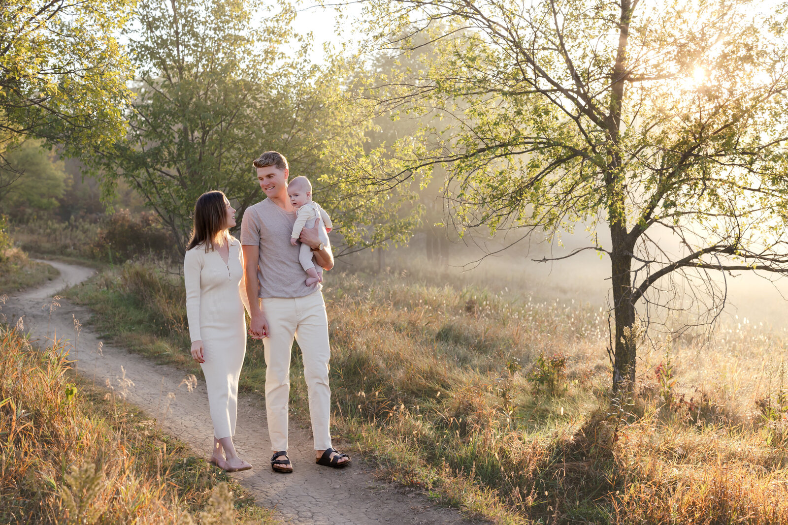 a family strolls through a narrow pathway in a the glowing sunrise light. The mom and dad are holding their small baby and are all dressed in neutral clothing