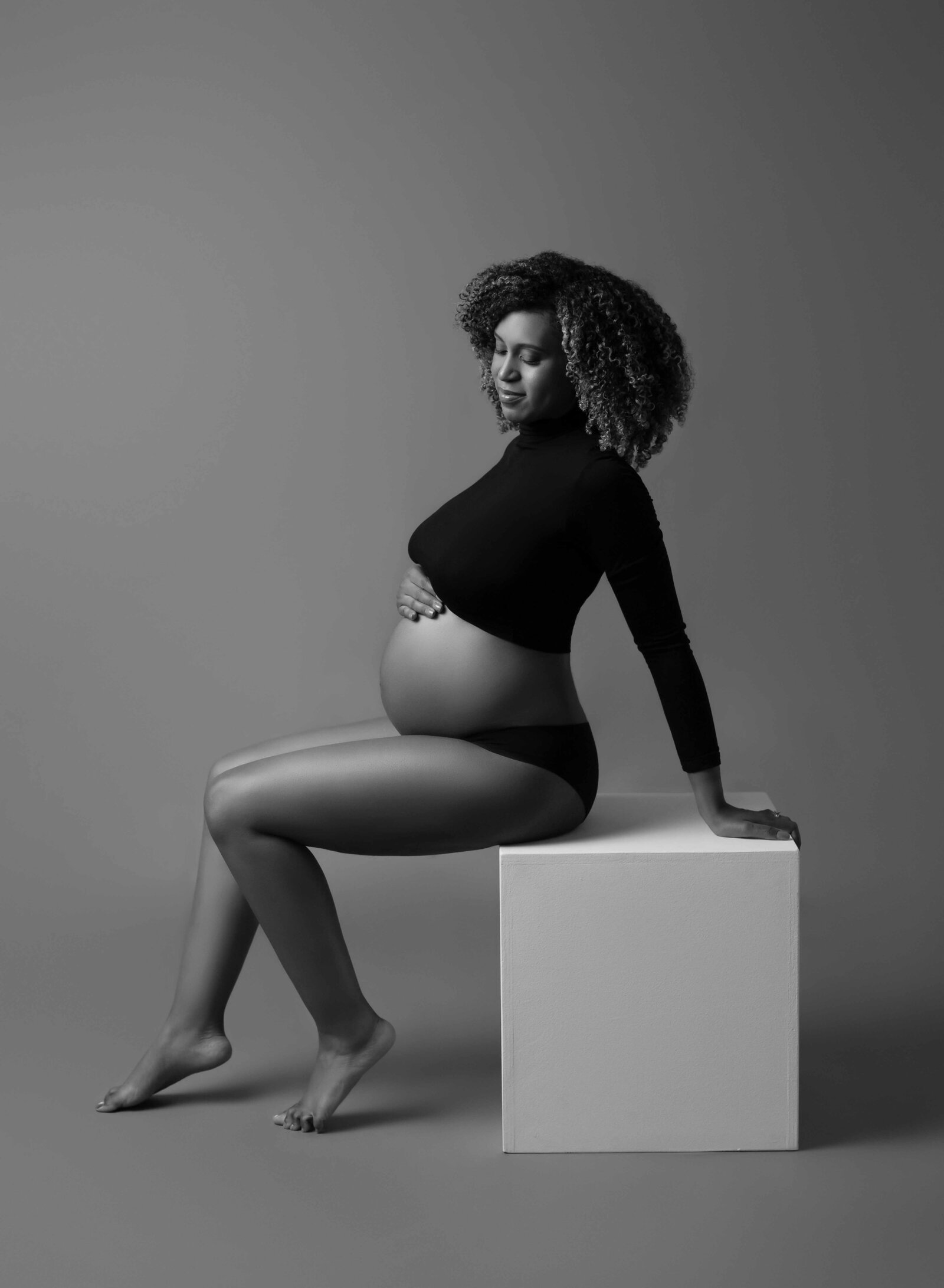 Pregnant women sitting on white box, looking at her baby bump. 