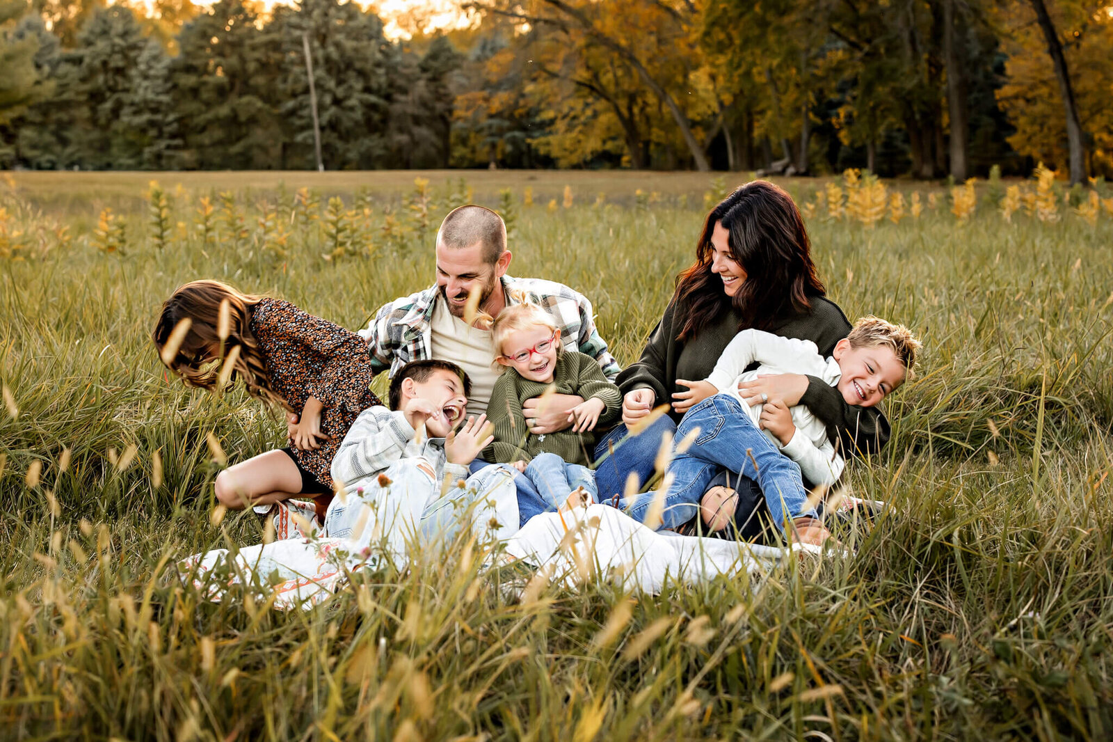 A Bloomington family photographer captures a joyful scene as a family sits on a blanket in a grassy field during sunset. The father, mother, and four children are laughing and playing together, surrounded by tall grass and trees. The mood is carefree and vibrant, perfect for outdoor portraits.