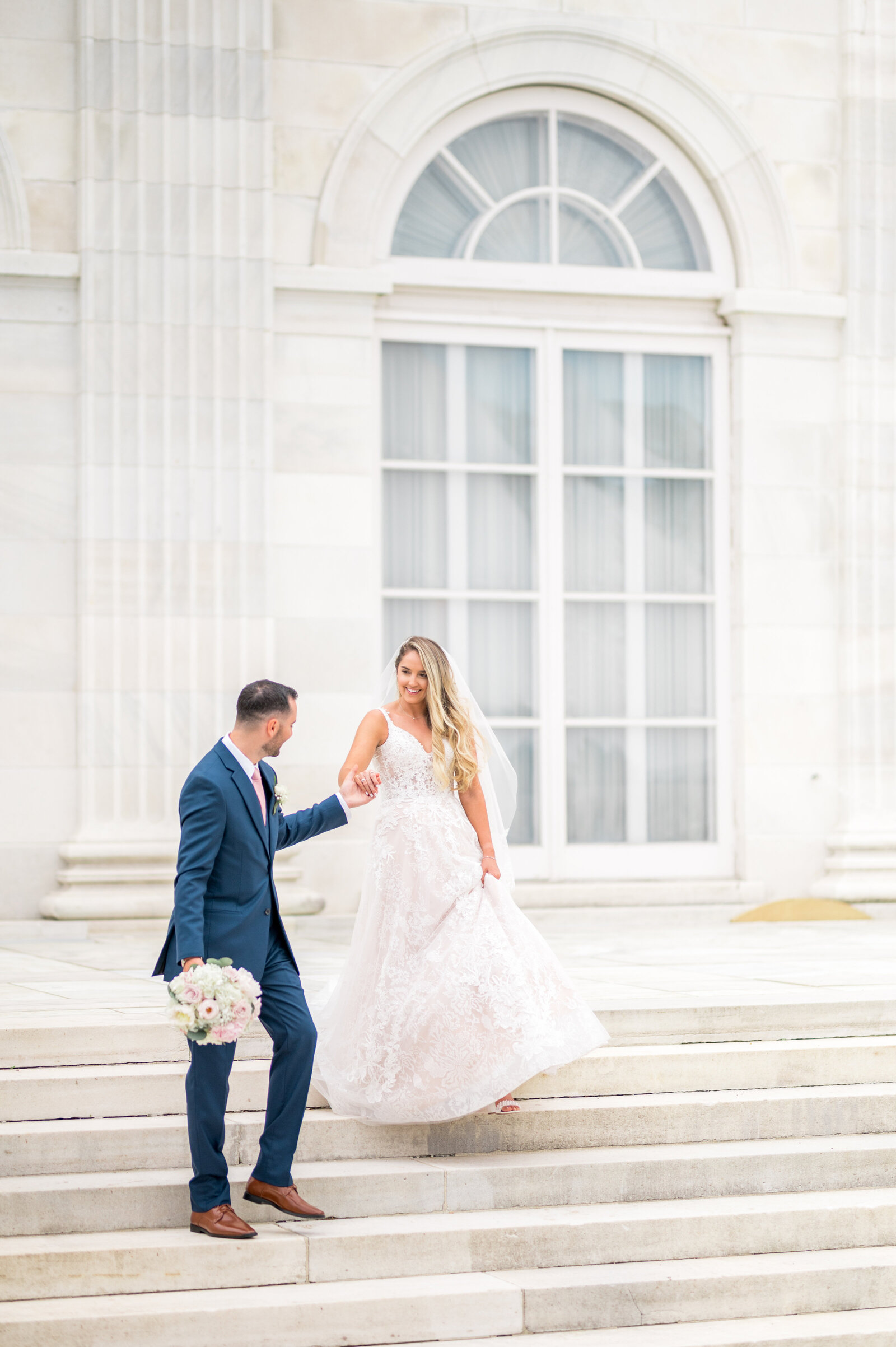 Bride gracefully descending classic stone staircase while groom reaches her hand, timeless and warm floor-to-ceiling window light.