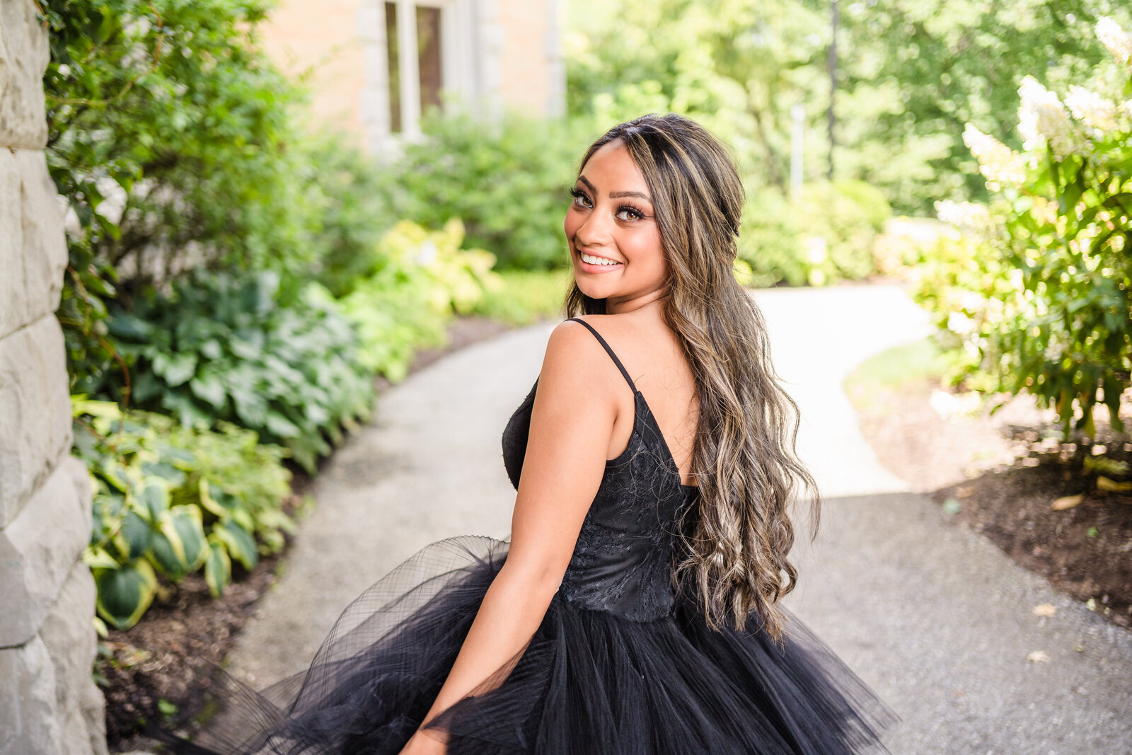 A bride in a black dress runs along a garden path and looks back at the camera smiling