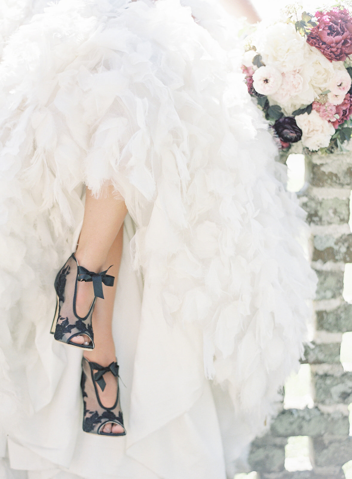 Black Bella Belle sheer high heel ankle boots with black floral detailing on toes and by heel. Open toed shoes and open on top of the foot. Tied together with a black satin ribbon in front. Bride's legs are crossed and all that is pictured is the open stone wall bride is sitting on, her white feathered skirt of her Anne Barge wedding gown, and her mid calves to toes. Photographed by wedding photographers in Charleston Amy Mulder Photography