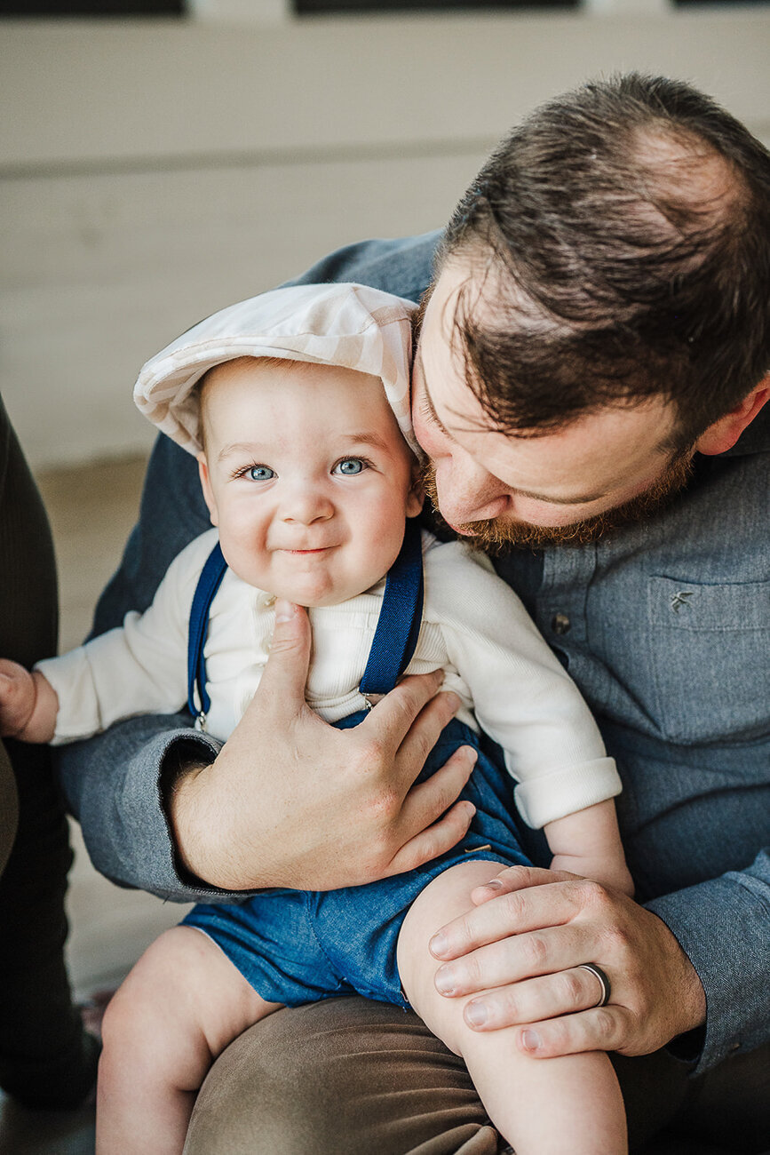 overhead shot of chubby baby in overalls staring into camera from dad's lap
