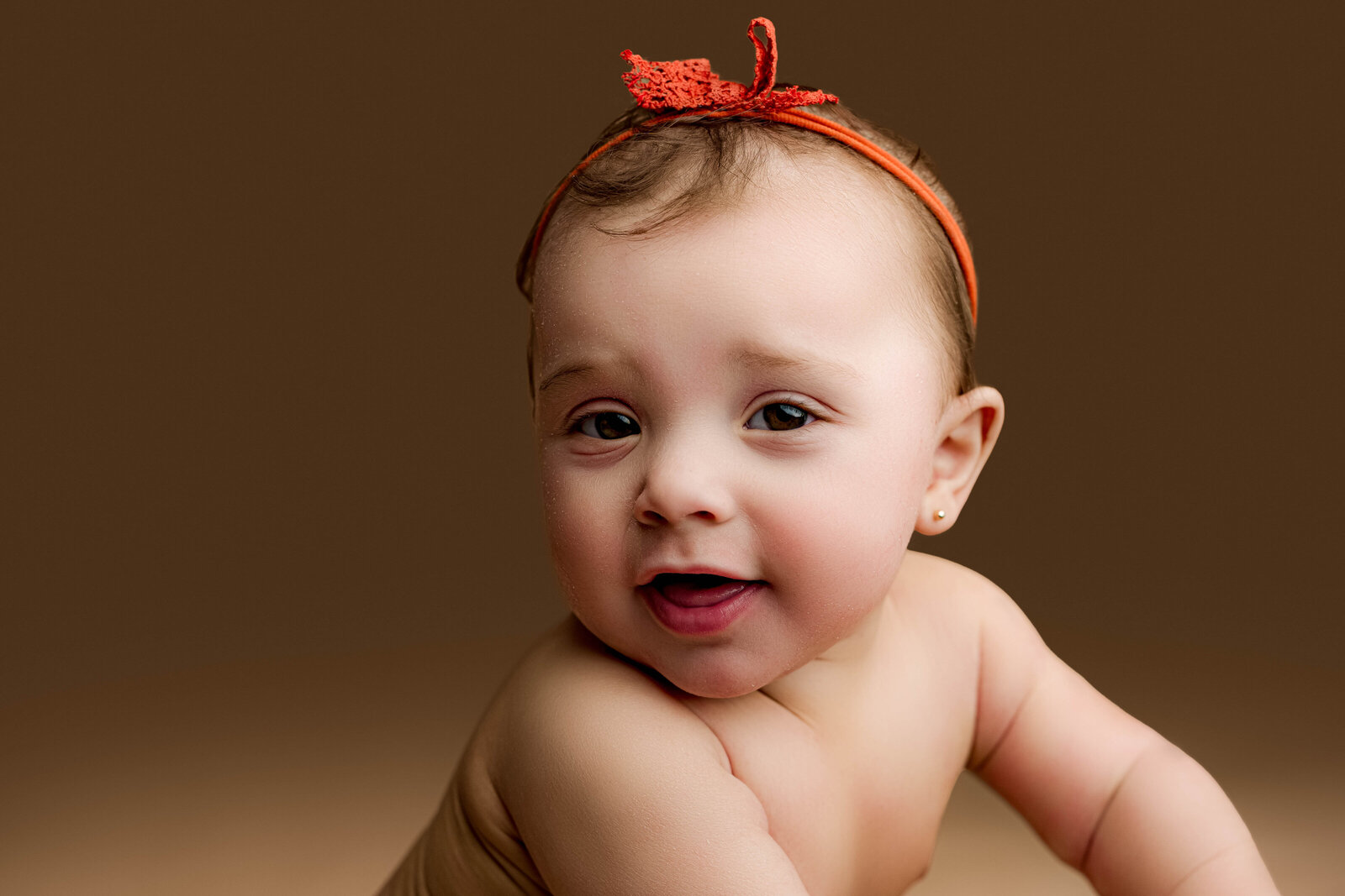 Close-up of a baby girl in an orange headband, smiling brightly as she leans toward the camera.