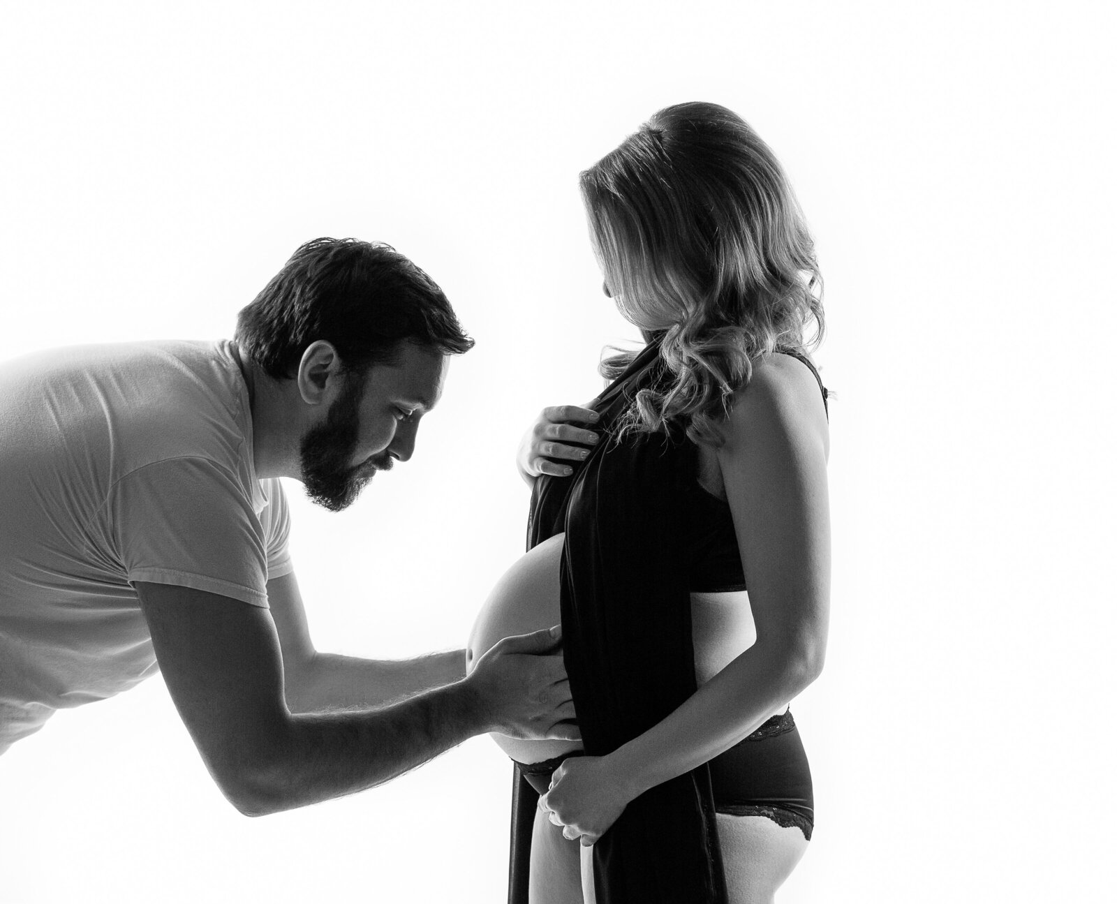Black and white studio maternity portrait on a white back ground of a pregnant Charlotte, NC couple, Mom to be's face is hidden behind her hair abut her husband is holding her bare belly . Dad has dark hair and is wearing a white t-shirt mom is draped in black fabric