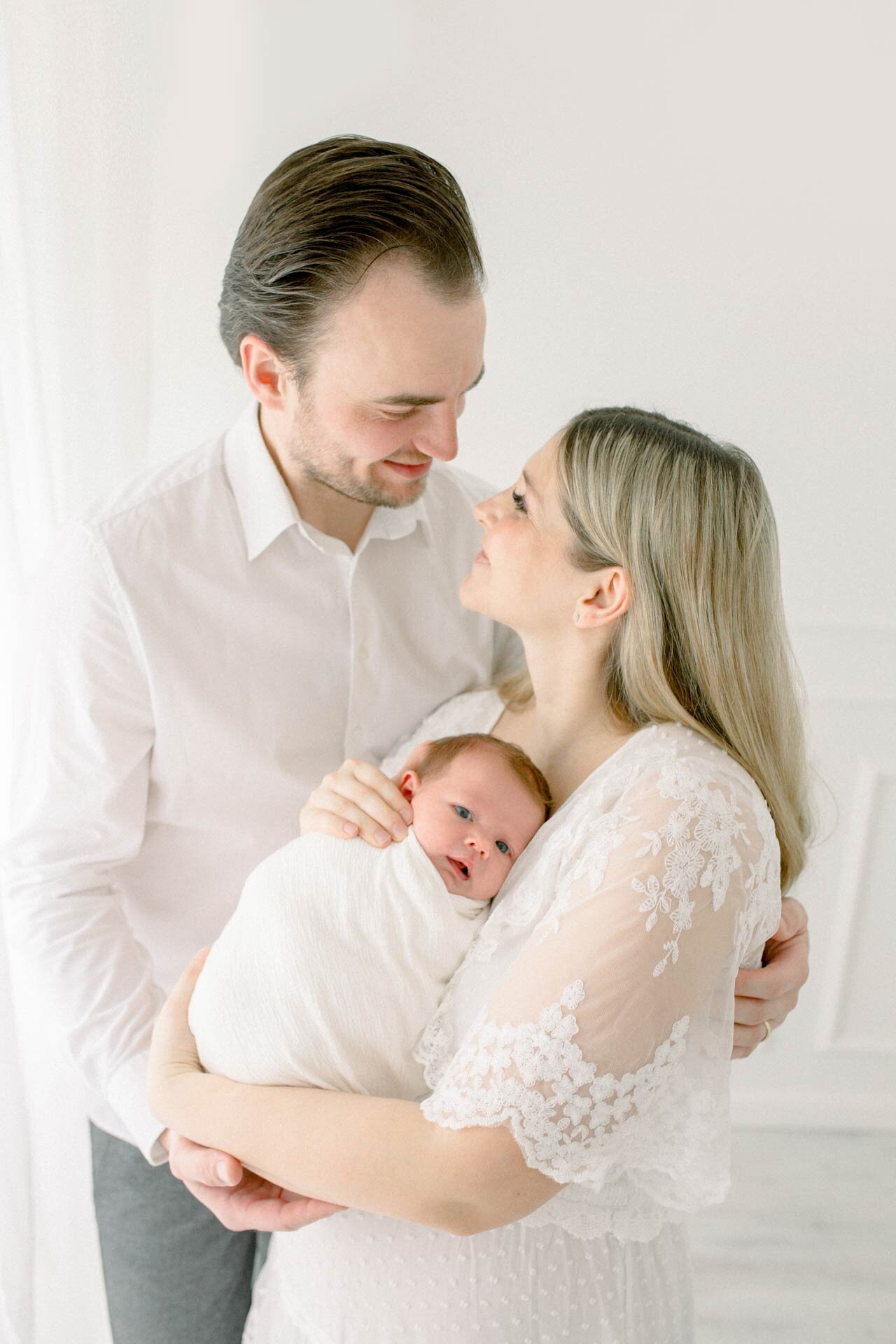 Neugeborenenfotoshooting nahe Bielefeld in weiß: Mutter in weißem Spitzenkleid hält Neugeborenes im Arm. Der Vater umarmt beide und Eltern sehen sich glücklich in die Augen.