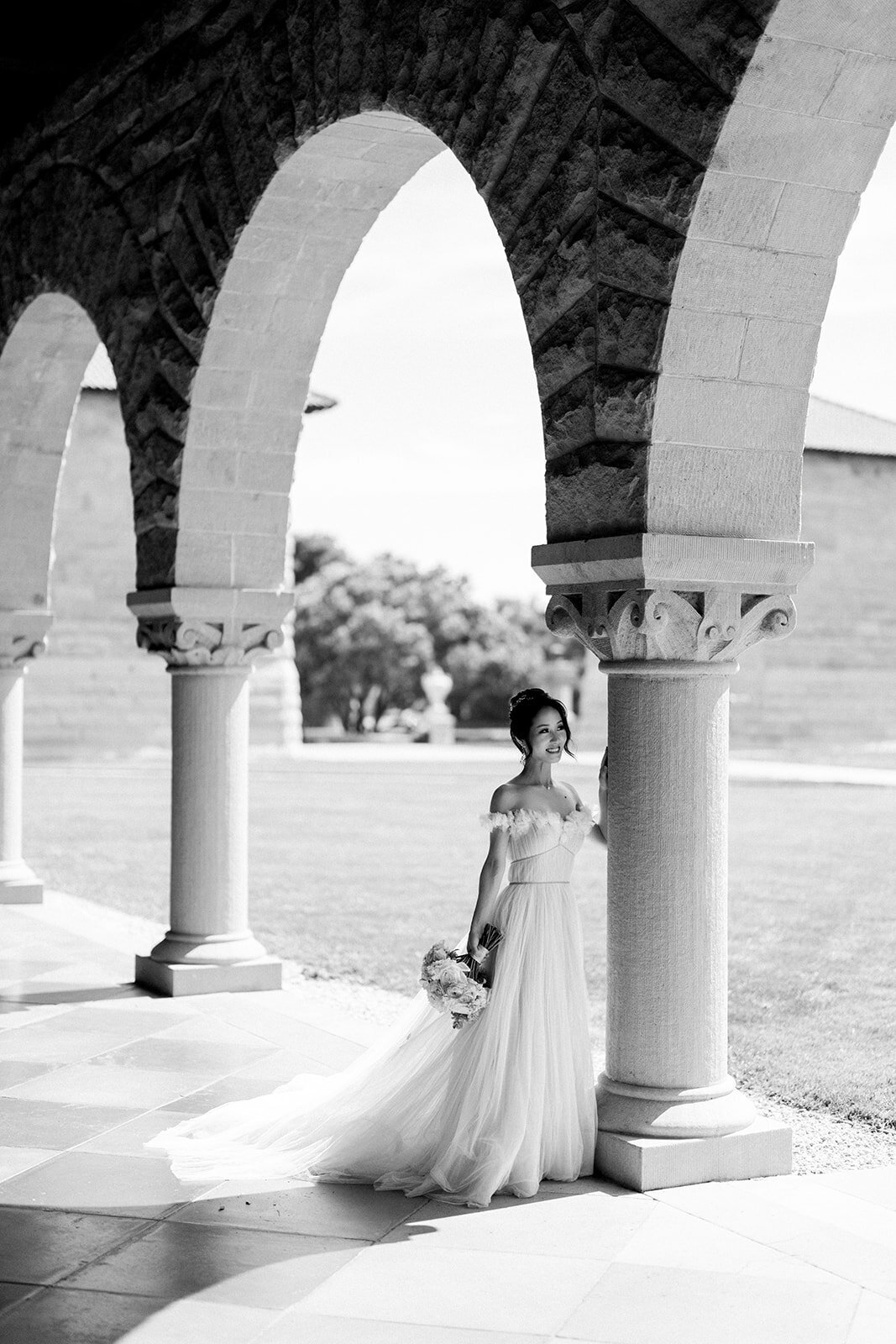 The bride stands elegantly by the archway at Stanford University, showcasing her graceful gown and serene beauty.