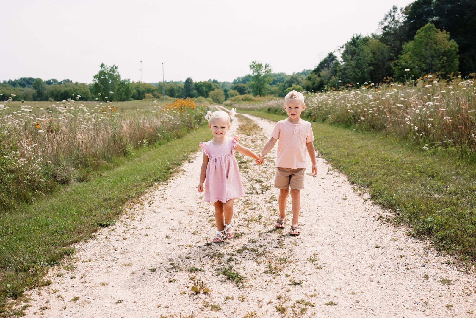 Happy toddler brother and sister both in pink walk hand in hand through a park trail lined with wildflowers