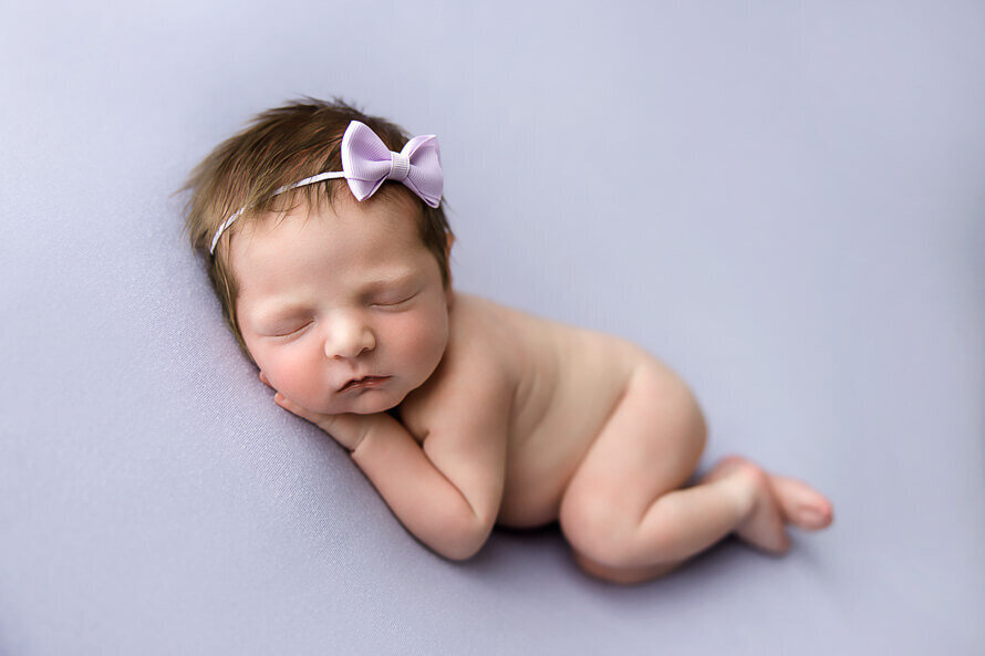 Newborn baby girl with a bow sleeps on a purple backdrop.