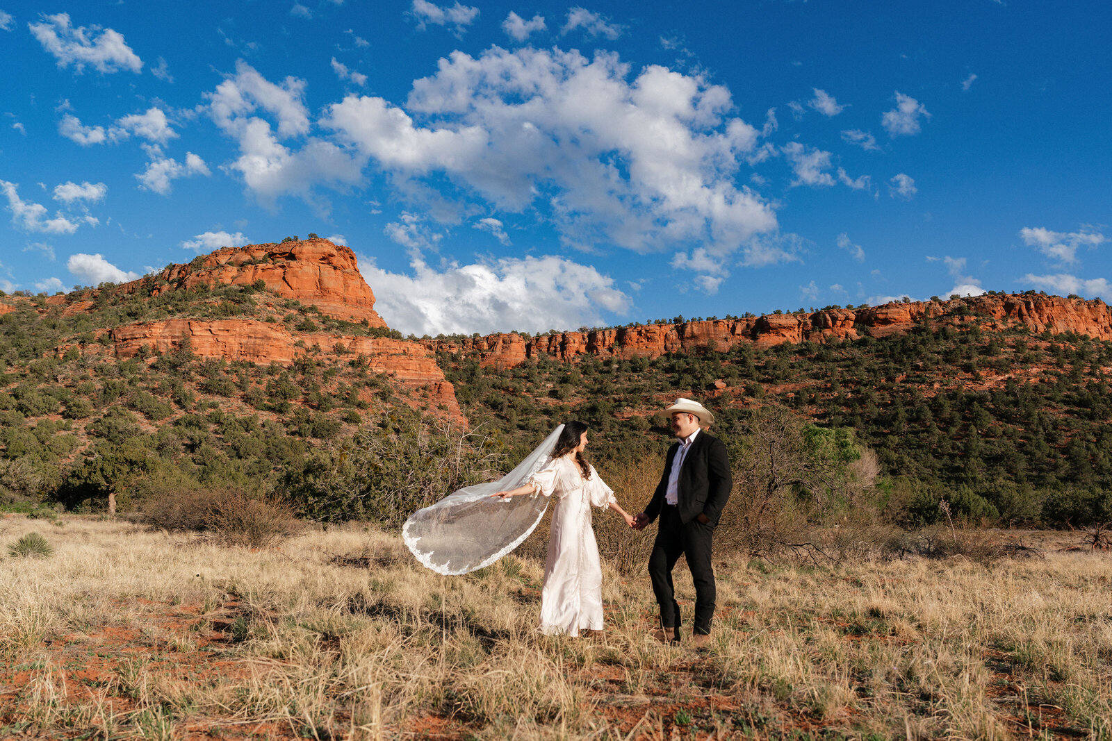 Bride and groom holding hands during their western-inspired elopement at Bear Mountain in Sedona, Arizona, with the bride's lace veil blowing in the wind against a backdrop of red rock cliffs and a vibrant blue sky.
