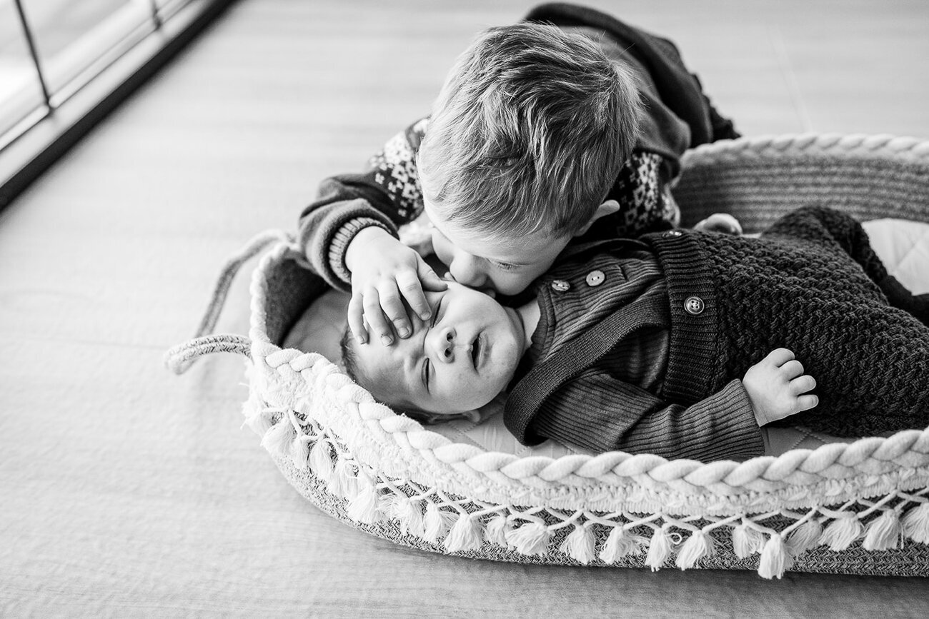 big brother leans over new baby in moses basket to kiss cheek