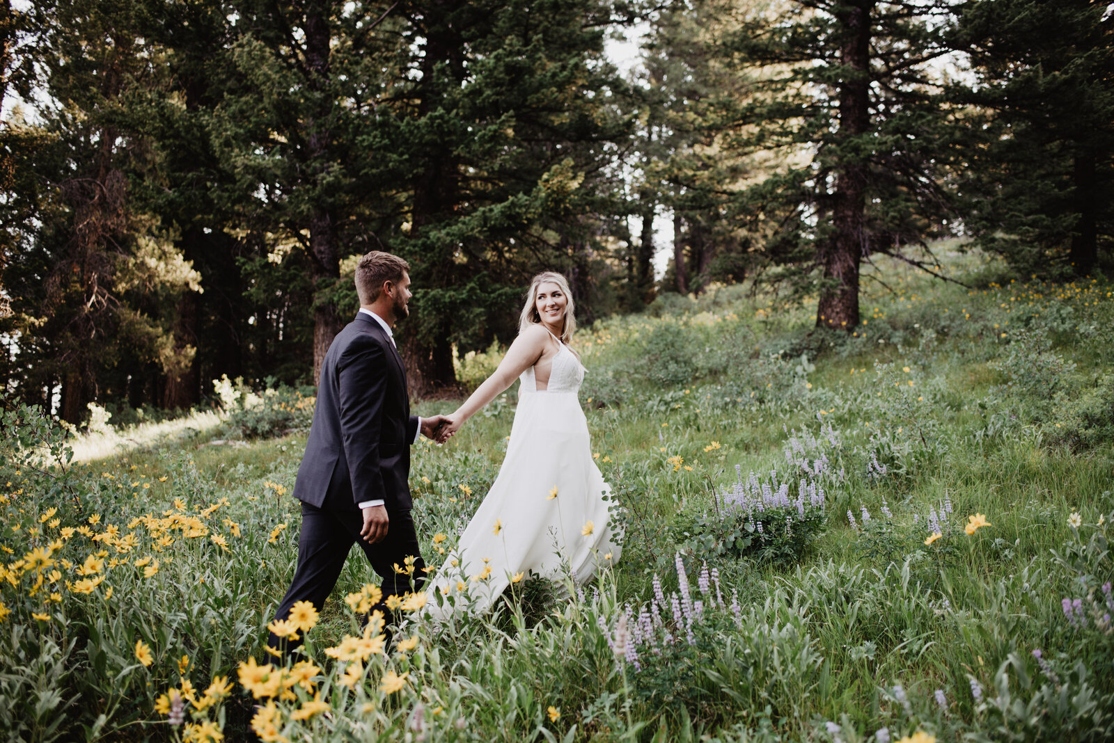 jackson hole photographers photographs bride and groom holding hands as the bride leads them up a hill in the woods with wildflowers