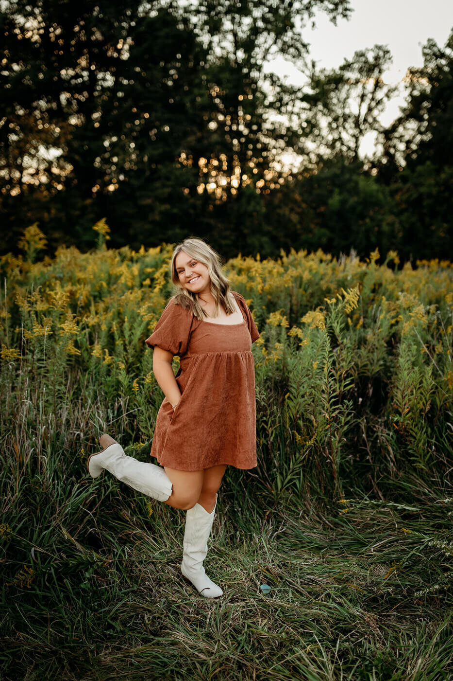 A person stands in a field with tall grass, wearing a brown dress and white boots. They are smiling with one leg bent back. The background includes trees and a slightly overcast sky.