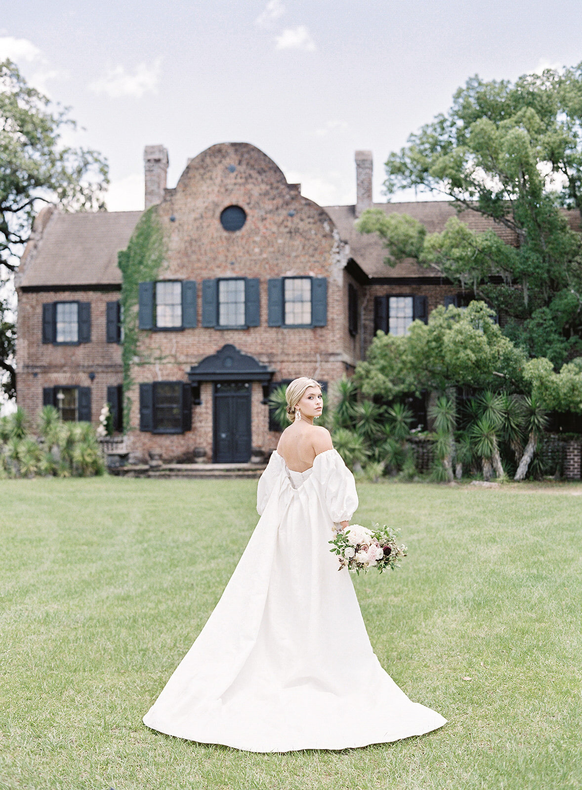 Bride in strapless wedding gown and ivory cape that is off the shoulders with a long train and has puffed sleeves designed by Anne Barge. The bride is facing away from the camera and looking back over her shoulder. Her pink, ivory, purple and taupe wedding bouquet is held down to her side in her right hand. She is in the front lawn of Middleton Place in Charleston South Carolina. The house is all brick, two stories and has black windows, shutters and front door.  Photographed by wedding photographers in Charleston Amy Mulder Photography.