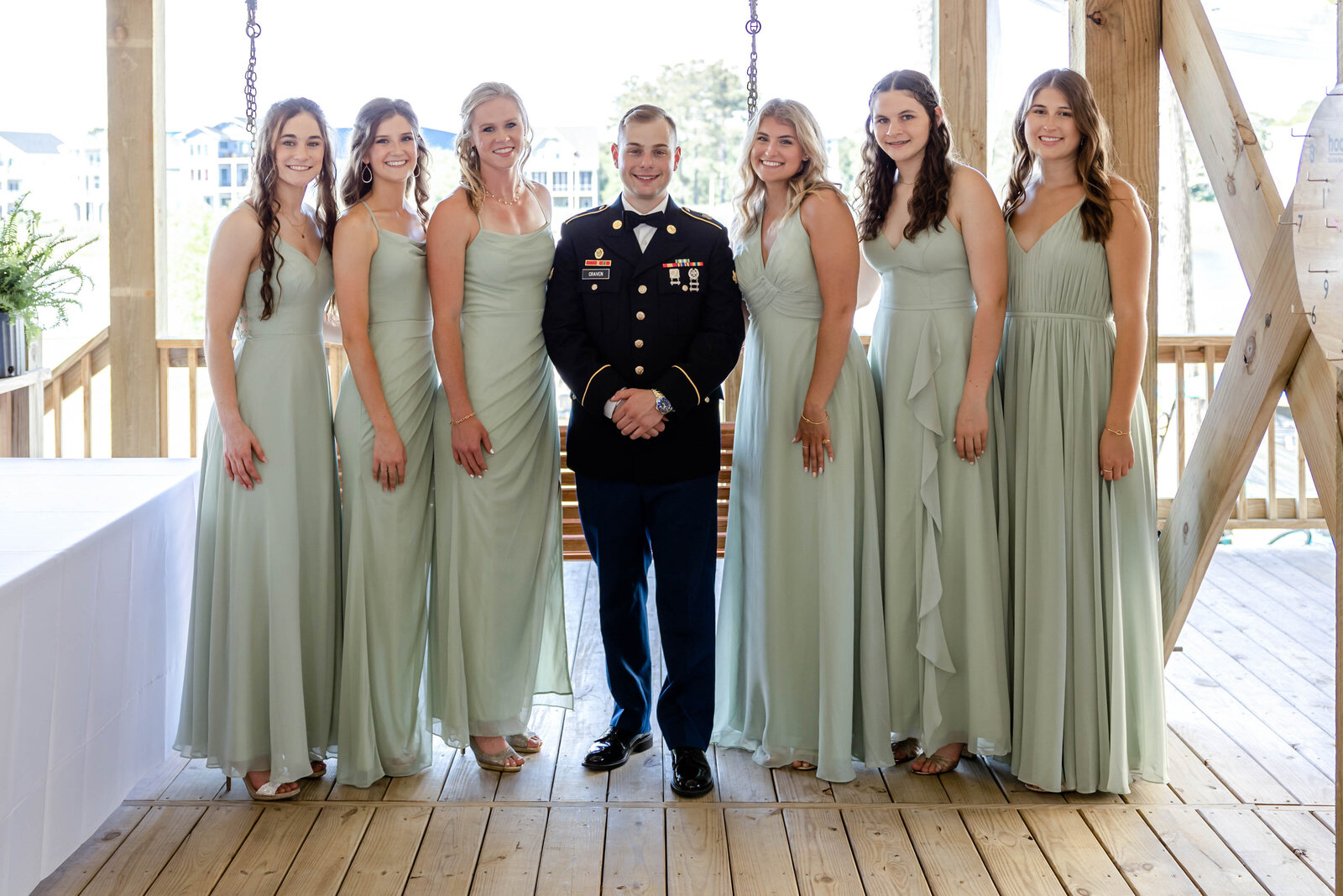 The groom, dressed in formal military attire, poses with bridesmaids in sage green dresses on a wooden deck at a Carolina Beach, NC wedding, blending coastal charm and timeless elegance.