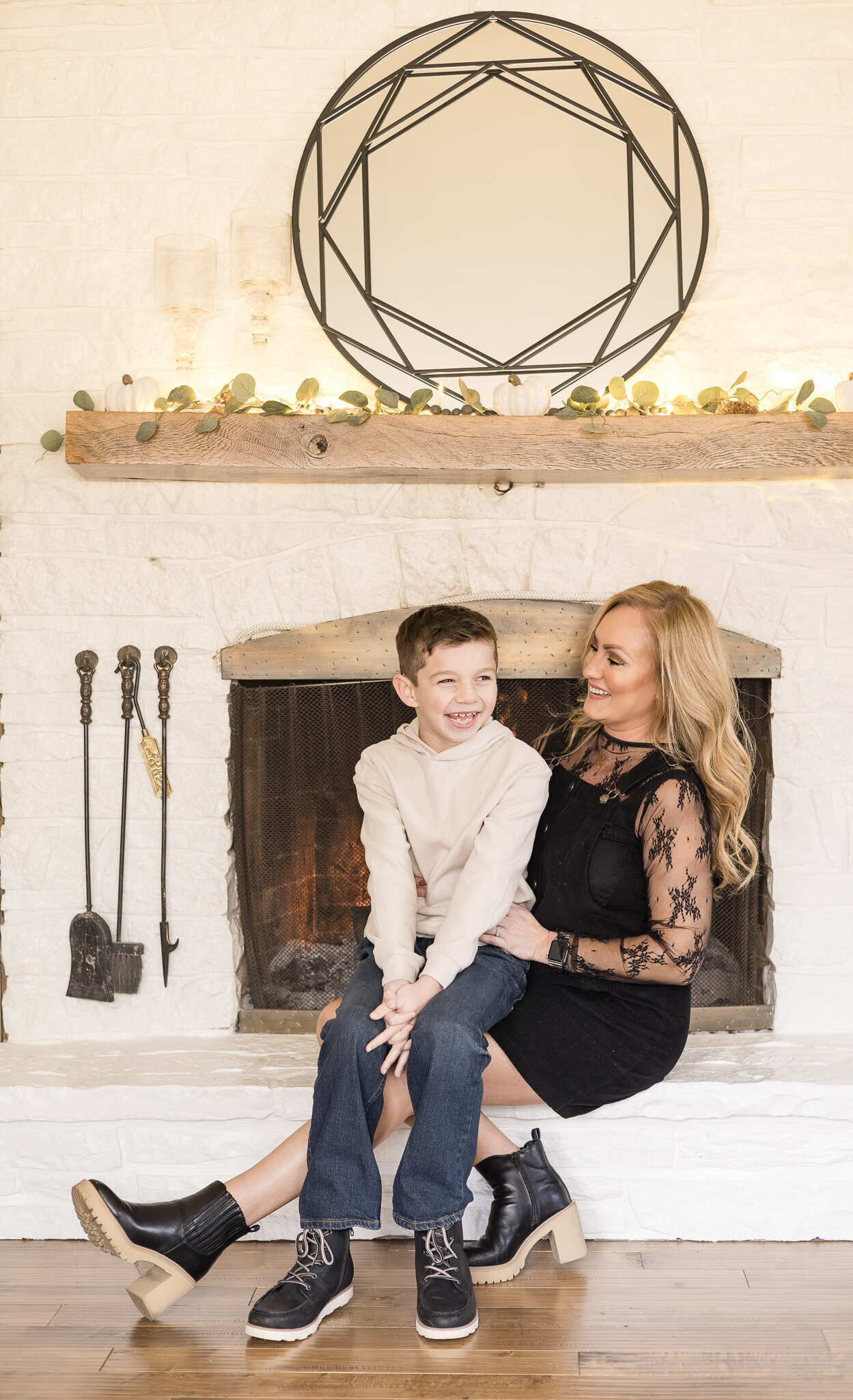 Mom and son sitting on the fireplace mantle. Mom has long blonde hair, wearing a black dress, looking at her son on her lap smiling.  Son has dark hair, wearing a long sleeve light tan shirt and blue jeans, laughing off of the camera