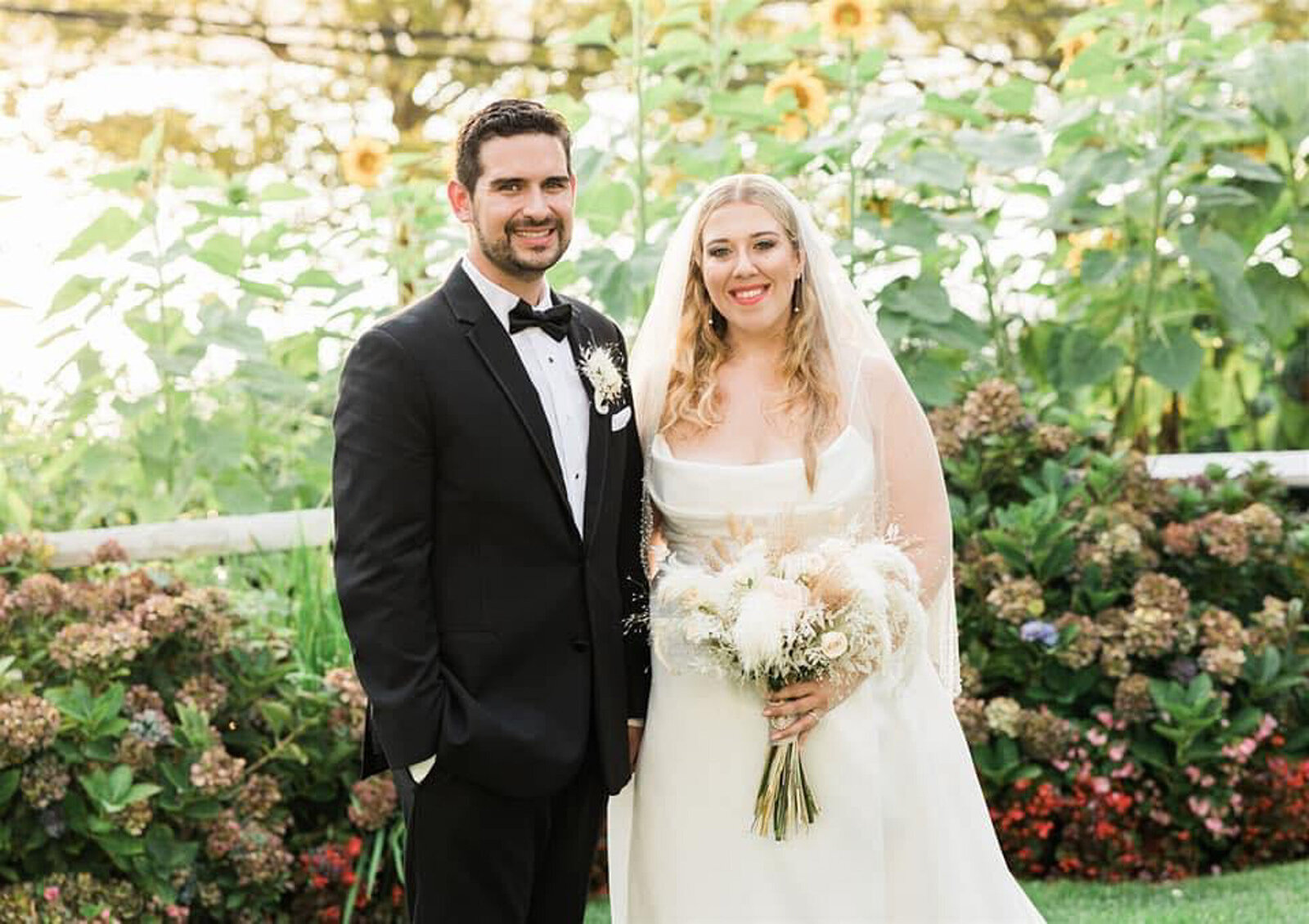 groom wearing a black tuxedo and the bride in a white gown holding a bouquet of flowers both smiling at the camera, with sunflowers and greenery in the background