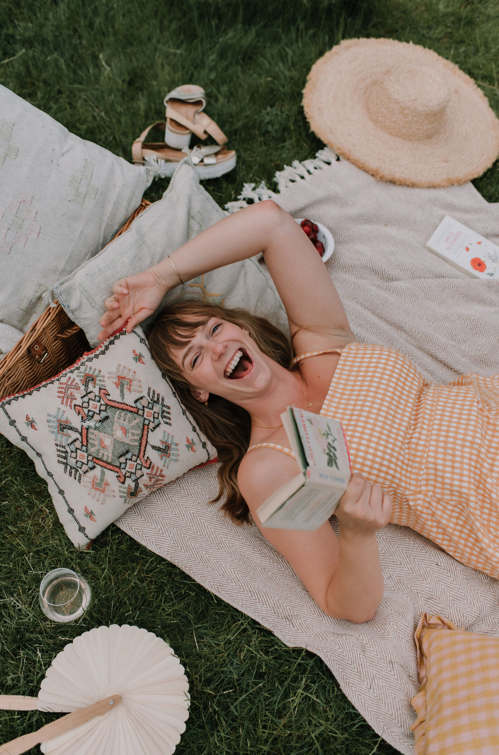 woman reading a book is laying on the grass surrounded by cushions and gingham patters, she is laughing