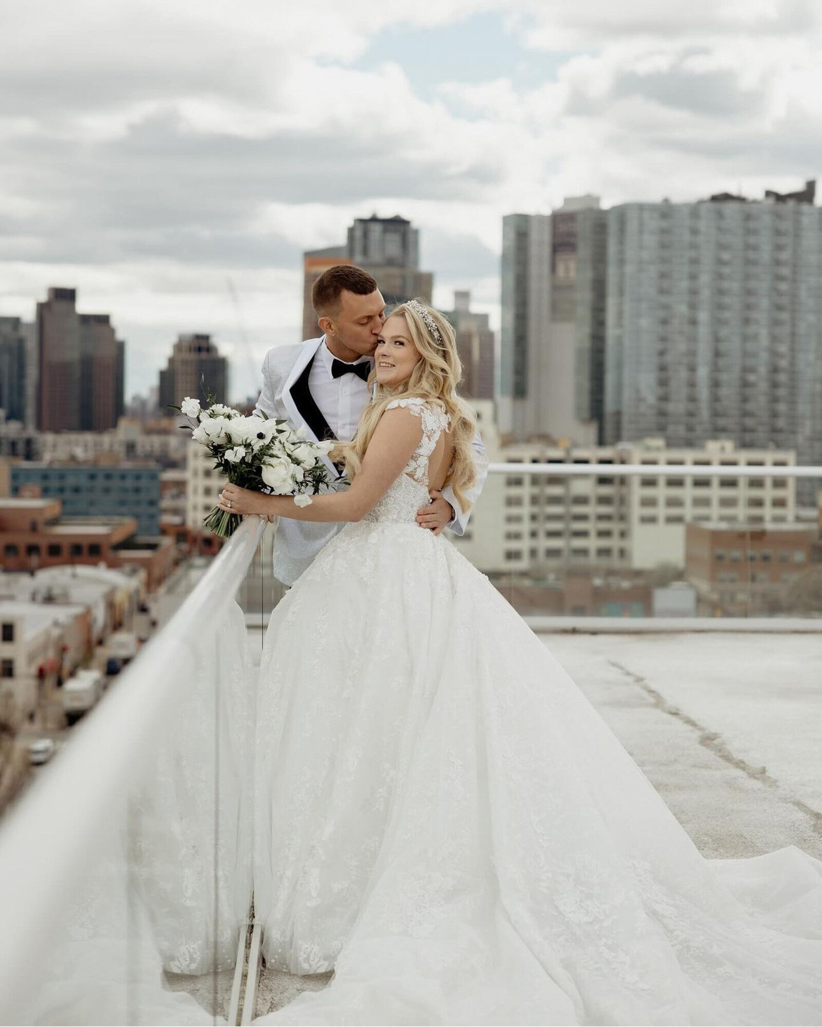 bride in a white lace wedding dress holding a bouquet of white flowers, while the groom is in a white tuxedo jacket, kissing her on the forehead both standing on a rooftop with a city skyline in the background