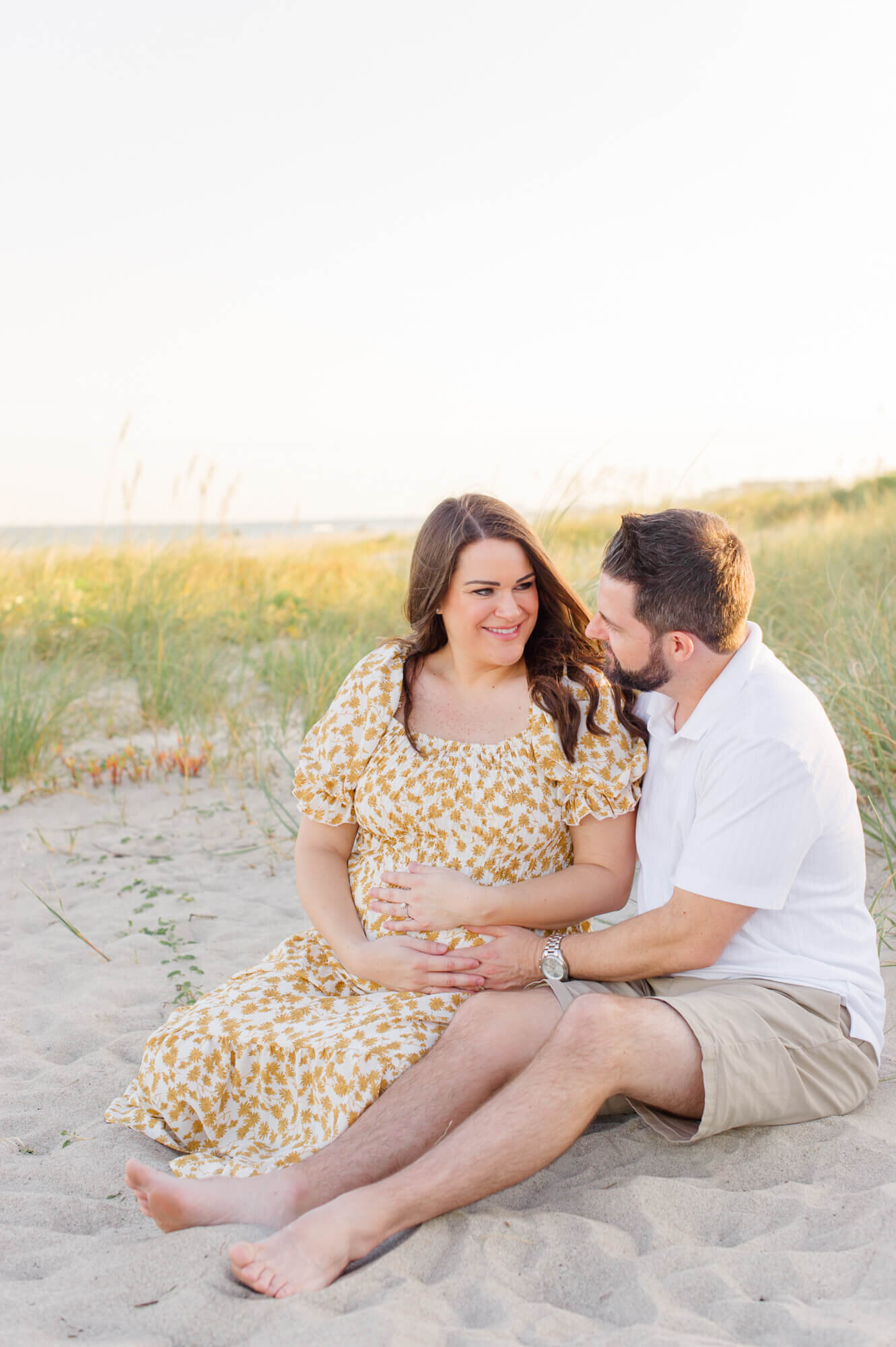New parents sit in the dunes at golden hour holding mothers belly and smiling at each other during their beach maternity session with an Orlando maternity photographer