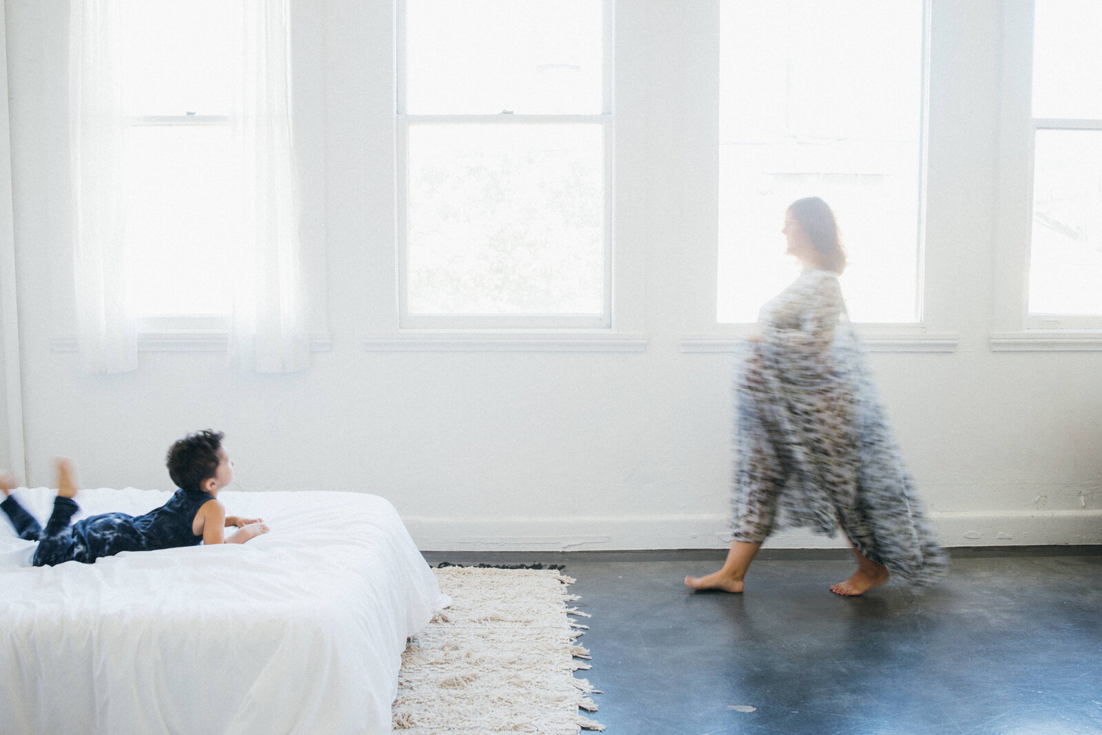 toddler laying on a white bed while mom in a flowing caftan walks towards him in front of large windows