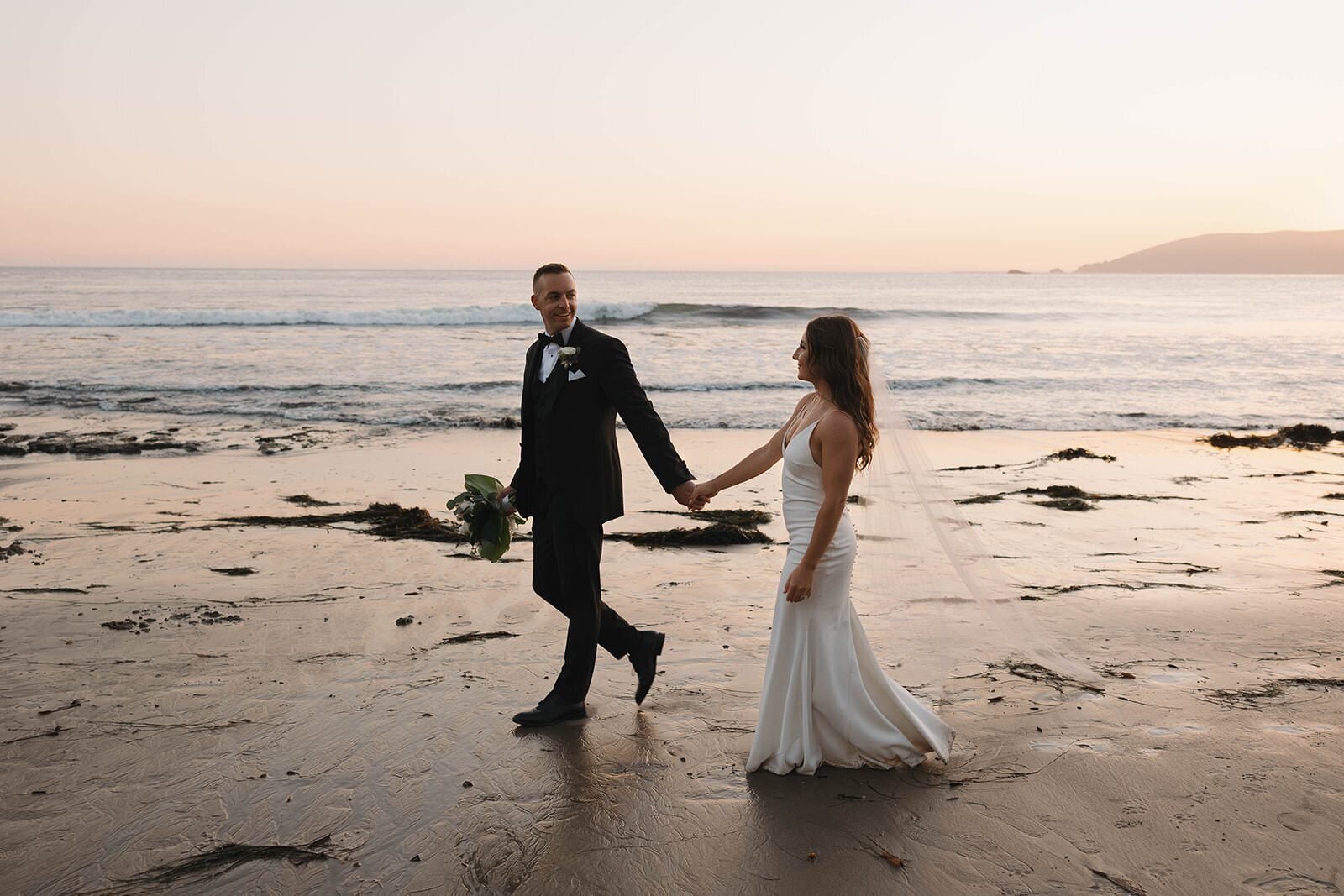 Bride and groom walking hand in hand along the beach at sunset